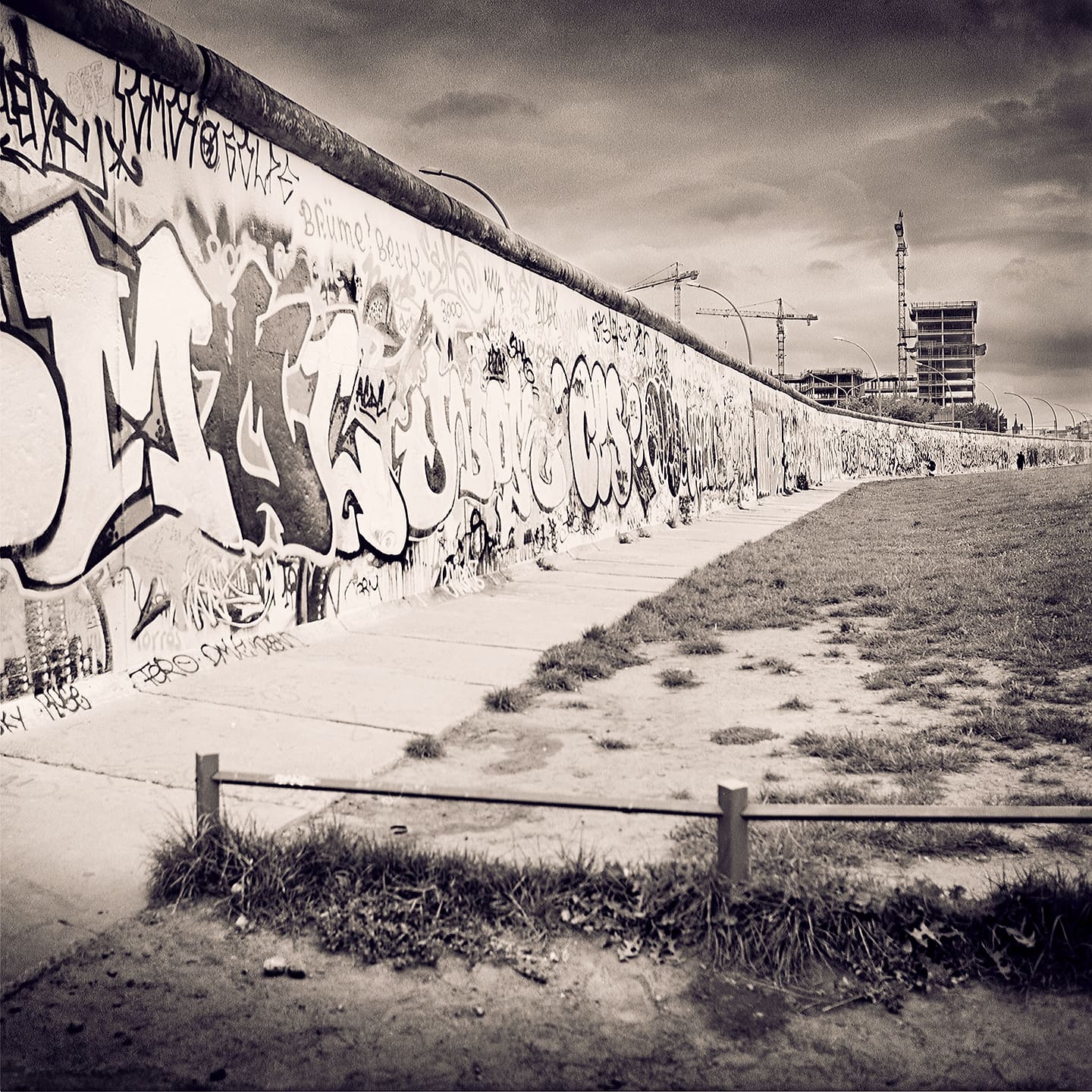 Graffiti-covered section of the Berlin Wall receding along a paved path, tower cranes over building work beyond