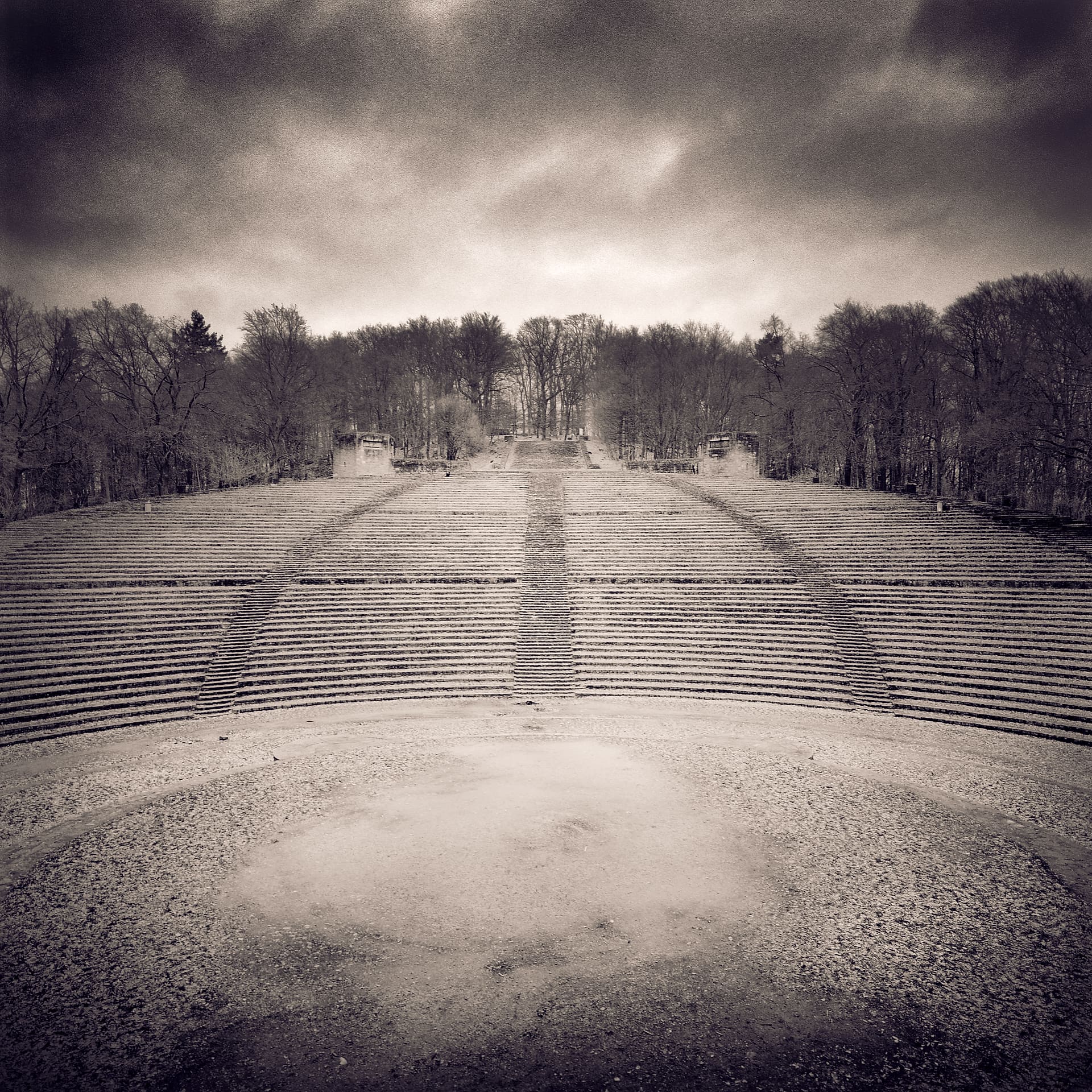 Empty open-air amphitheatre of stone terraces rising symmetrically to bare woods under heavy clouds