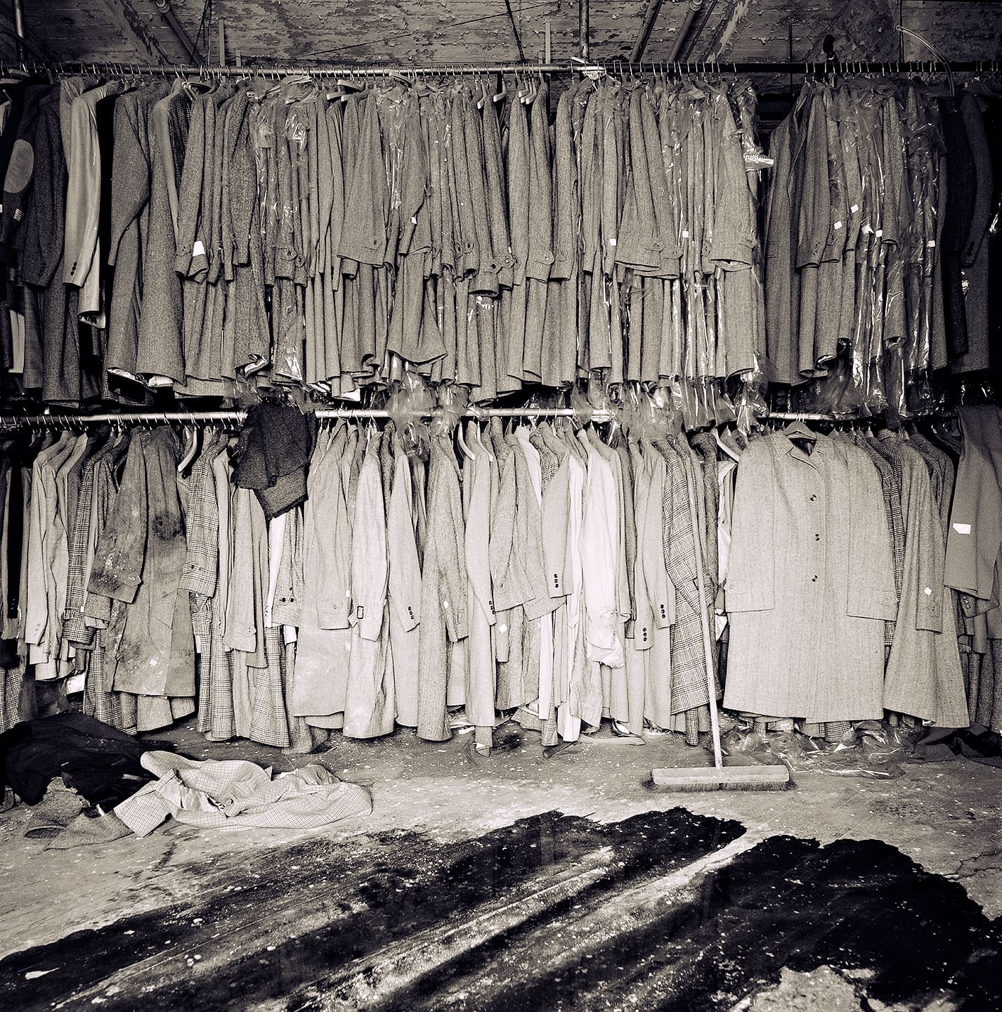 Racks of coats and suits still hanging in plastic in a derelict factory, debris and a broom on the stained floor