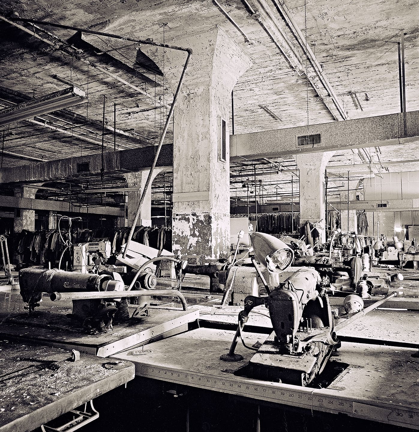 Rows of derelict industrial sewing machines and cutting tables under a peeling concrete ceiling, garments hanging behind