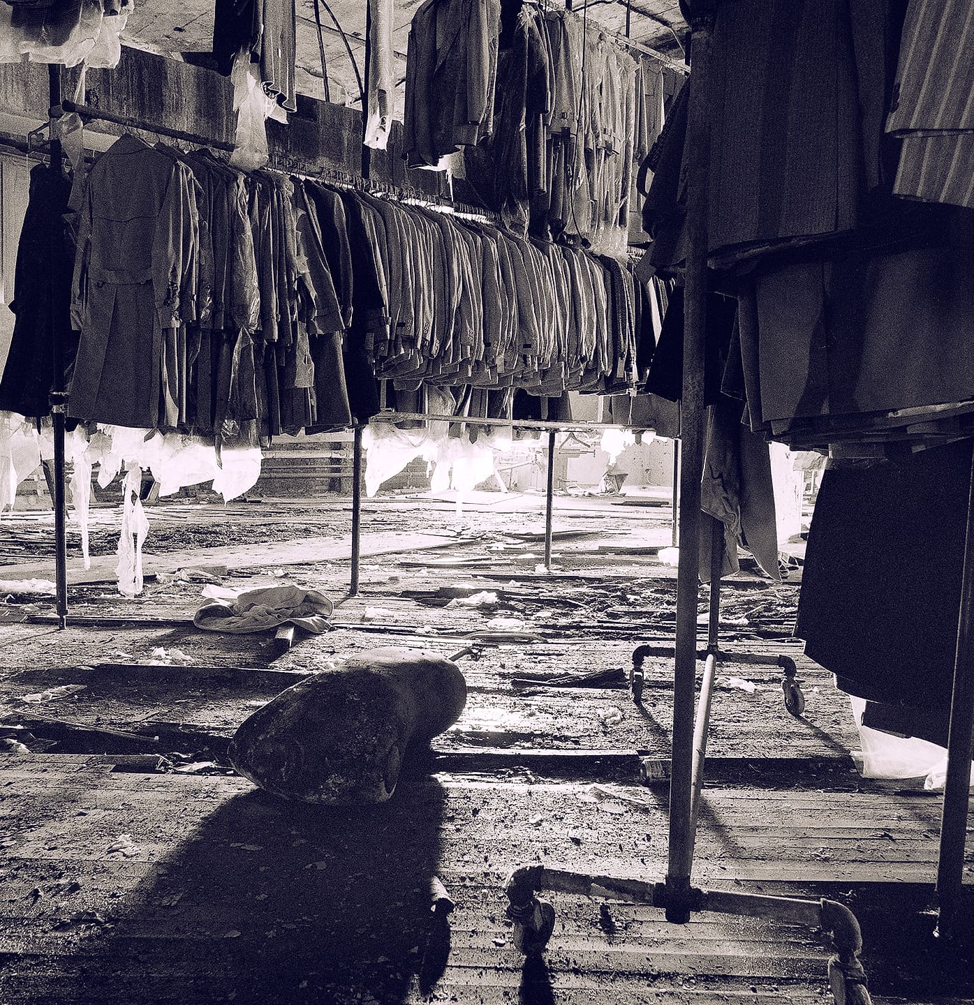 Low view along sagging garment rails hung with coats in a wrecked factory, torn plastic, rubble and daylight beyond
