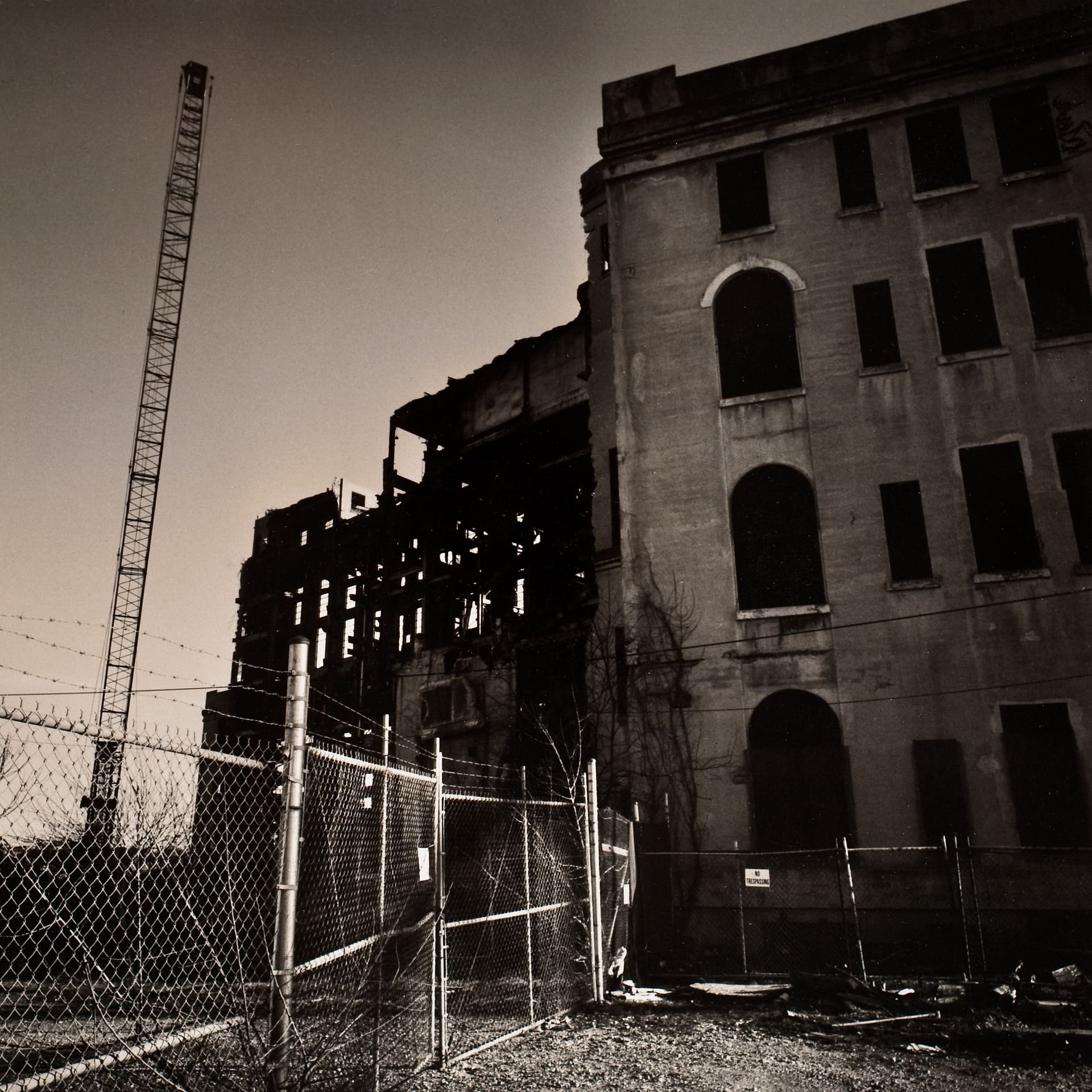 Gutted masonry power station behind chain-link and barbed-wire fencing, a lattice crane rising against a bare sky