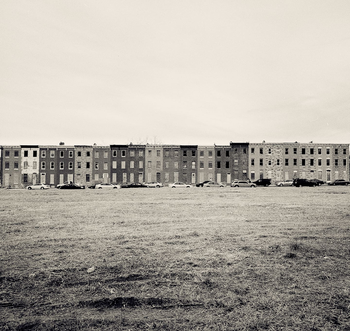 Long row of boarded-up brick rowhouses across a wide empty field of dead grass, parked cars along the street
