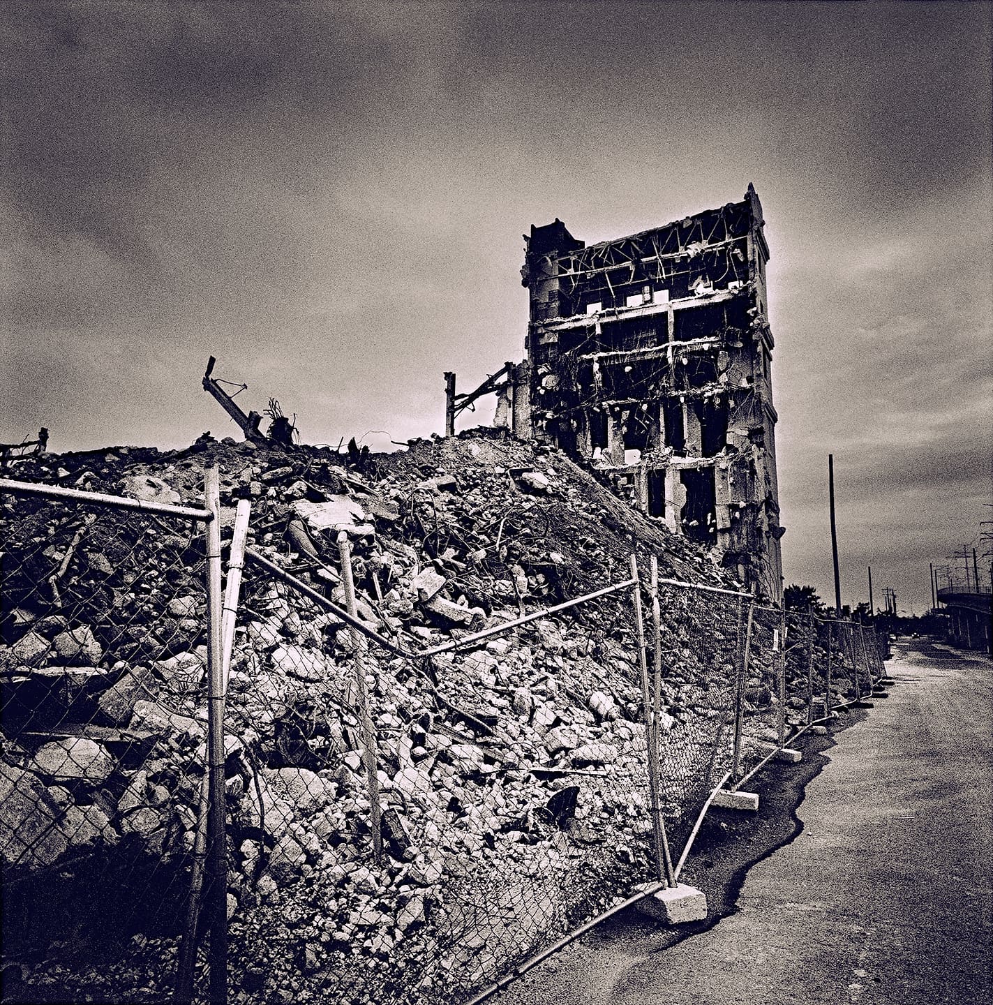 Half-demolished concrete tower stripped to its frame above a long rubble mound behind chain-link fencing beside an empty road