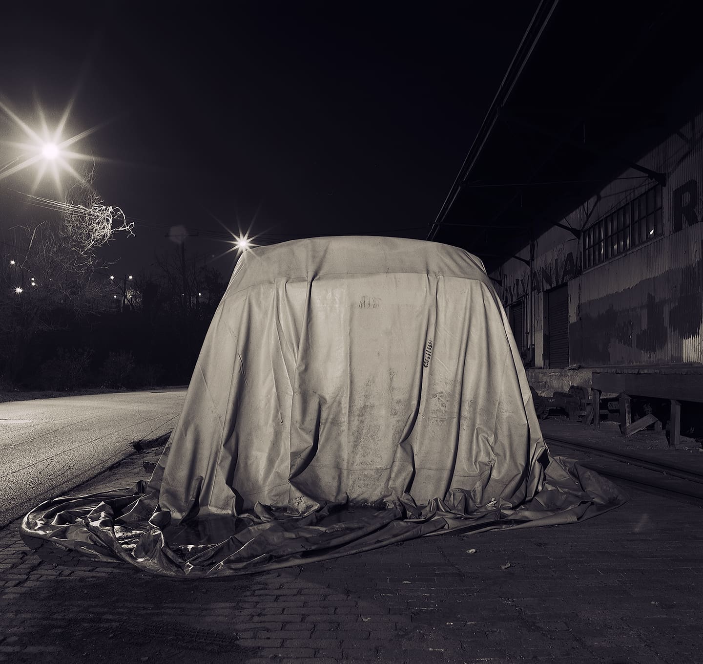 Large tarpaulin-shrouded object beside railway tracks and a corrugated shed at night, streetlamps flaring behind