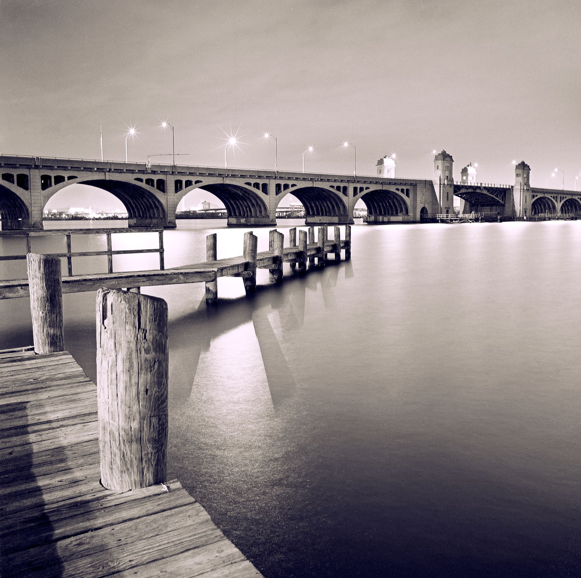 Arched concrete bridge lit at night above water smoothed to glass by long exposure, wooden pier pilings foreground
