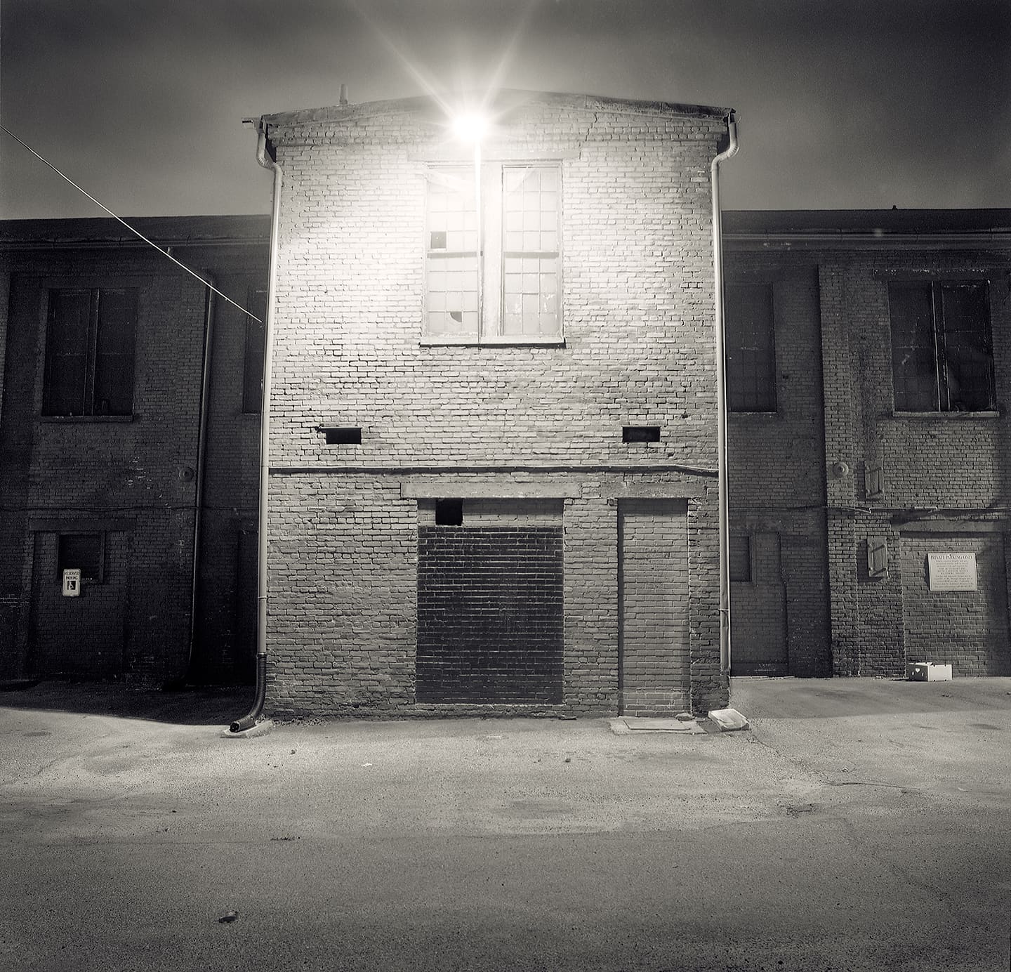 Small brick industrial building lit at night by a single flaring lamp above a broken upper window, bricked-up doorway below