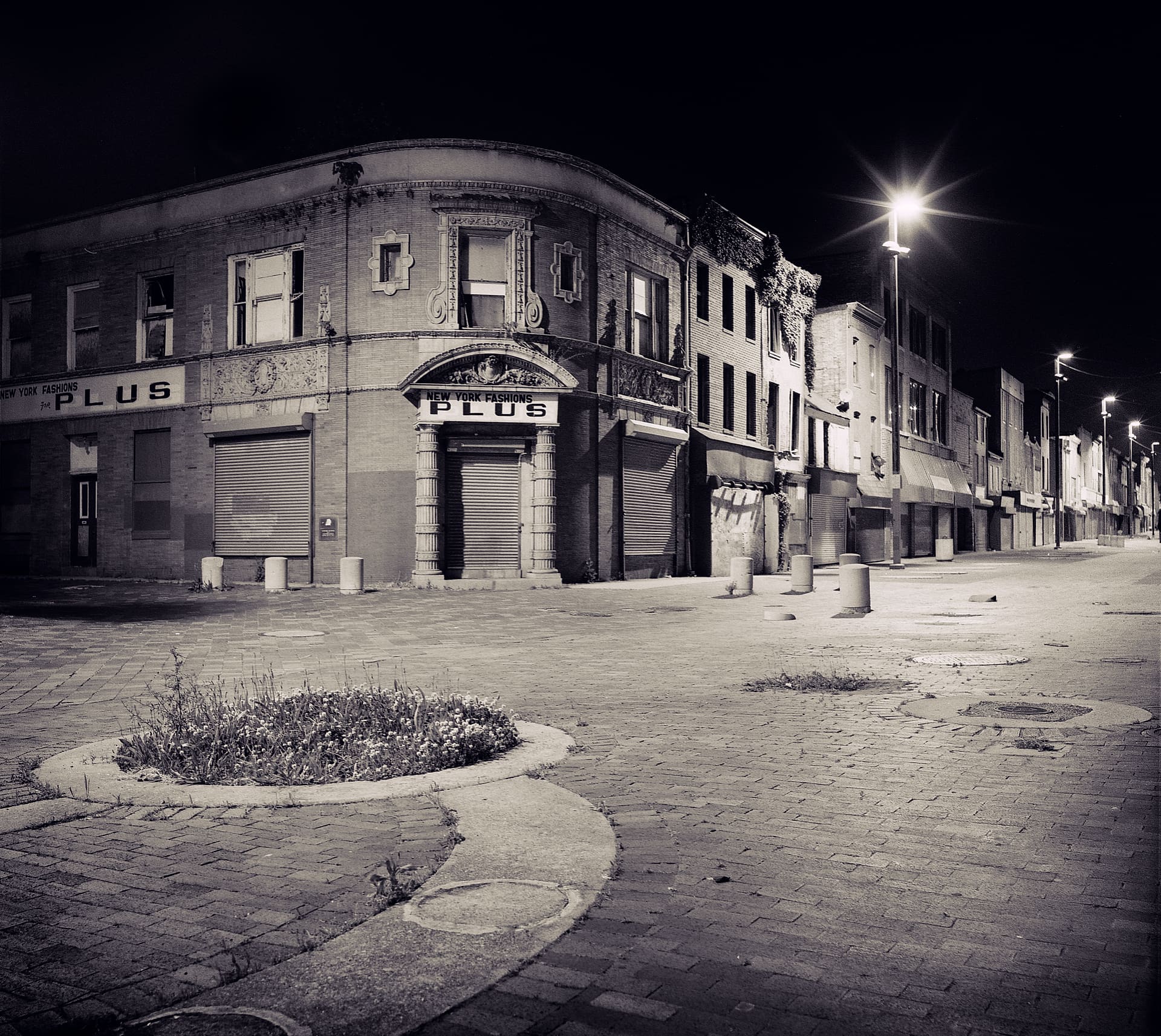 Empty brick pedestrian street at night, shuttered corner shopfront lettered New York Fashions Plus, weeds in a planter