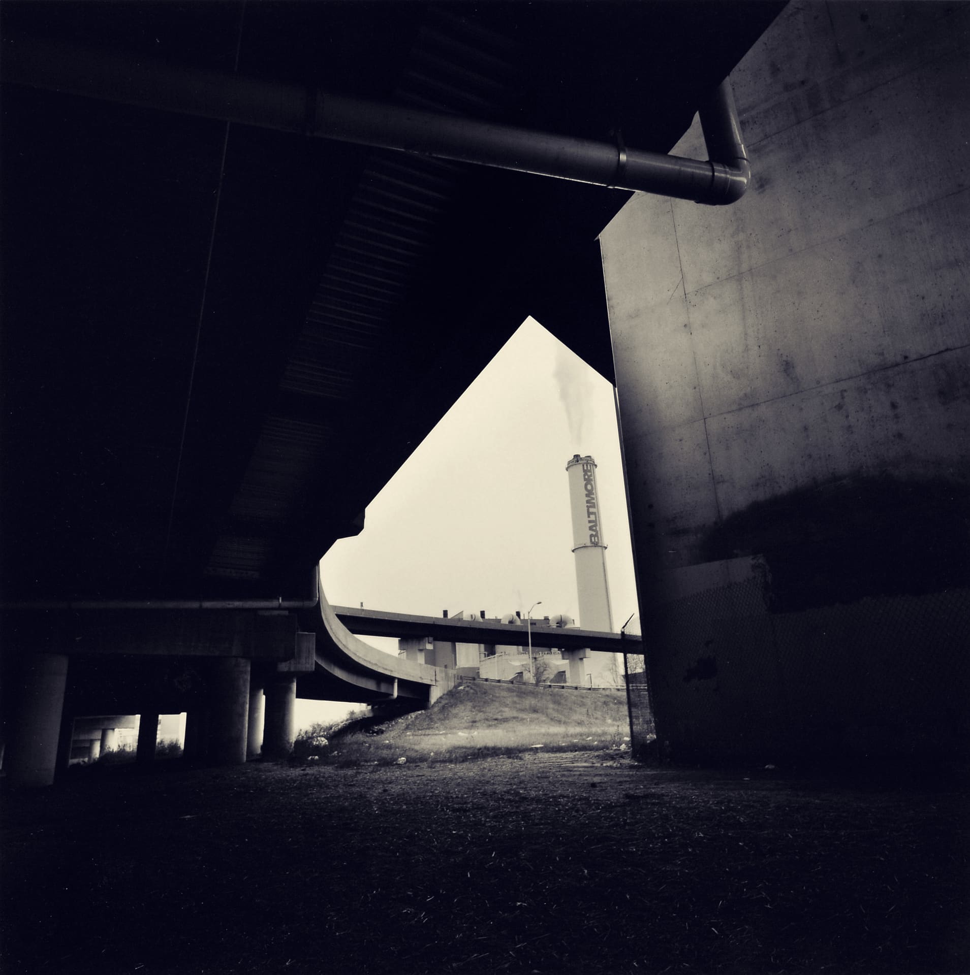 Dark underside of stacked highway ramps framing a triangle of pale sky and a smoking Baltimore-lettered chimney