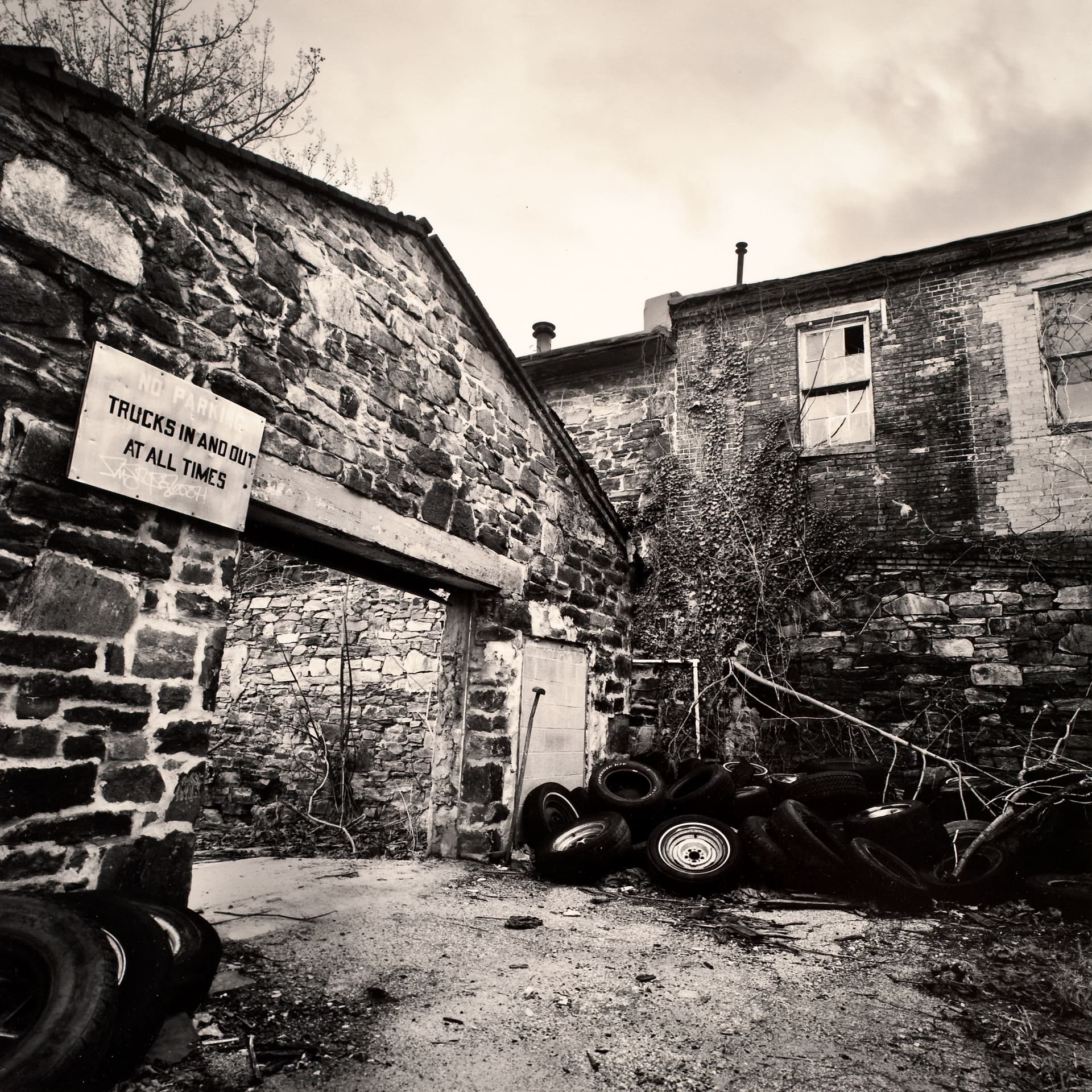Stone mill yard with a No Parking sign over a blocked archway, ivy-covered brick wall and dumped car tyres
