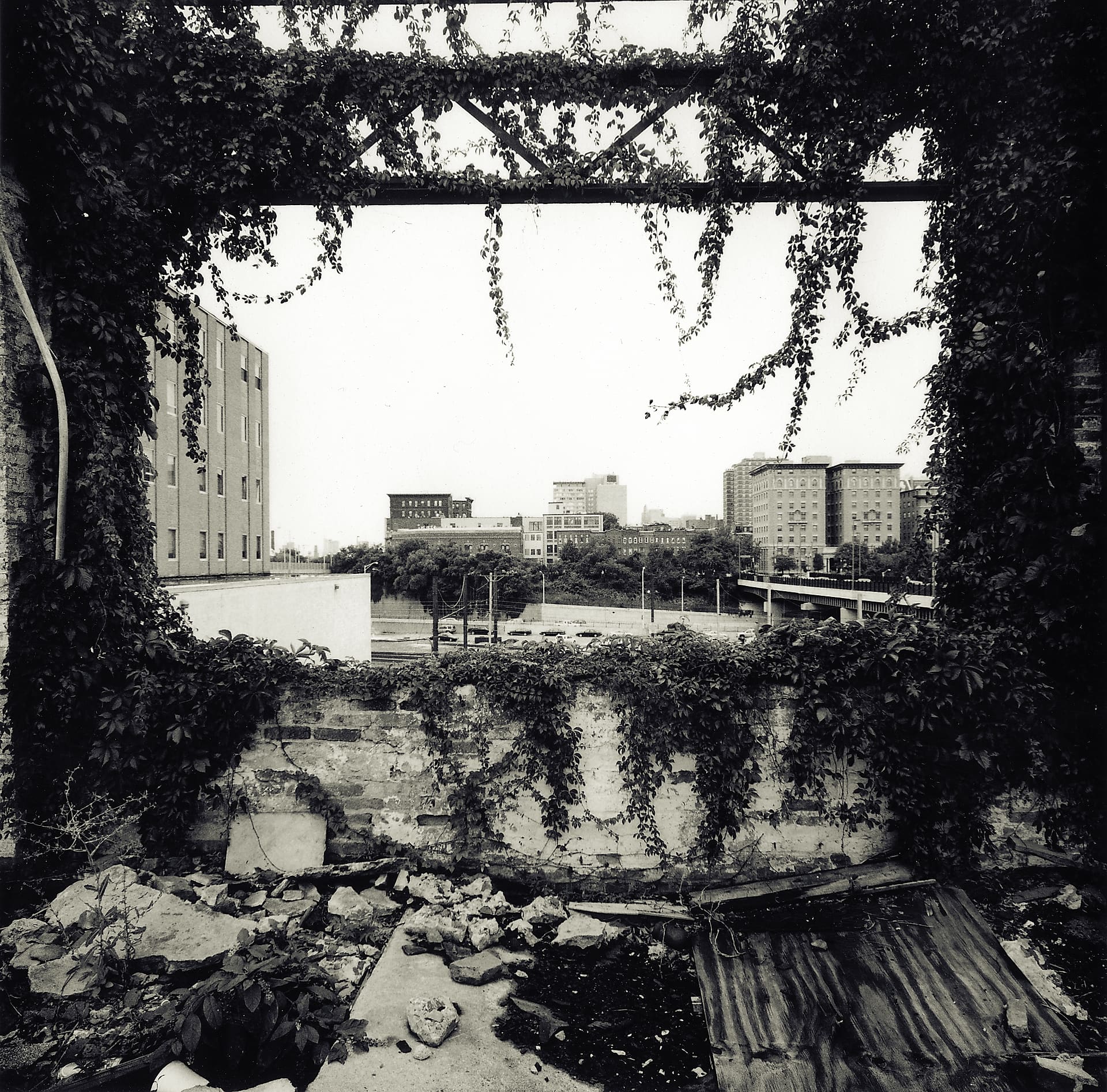 City skyline and elevated road framed through a ruined ivy-covered wall opening, rubble and rusted sheeting inside