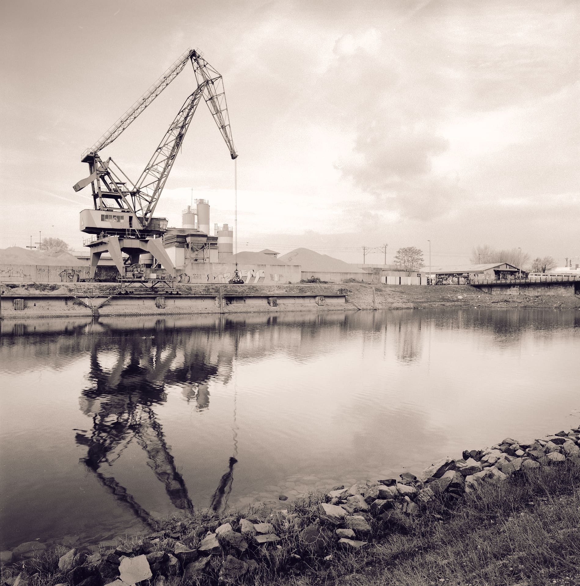 Disused harbour gantry crane and cement silos mirrored in still dock water, rip-rap stones along the near bank