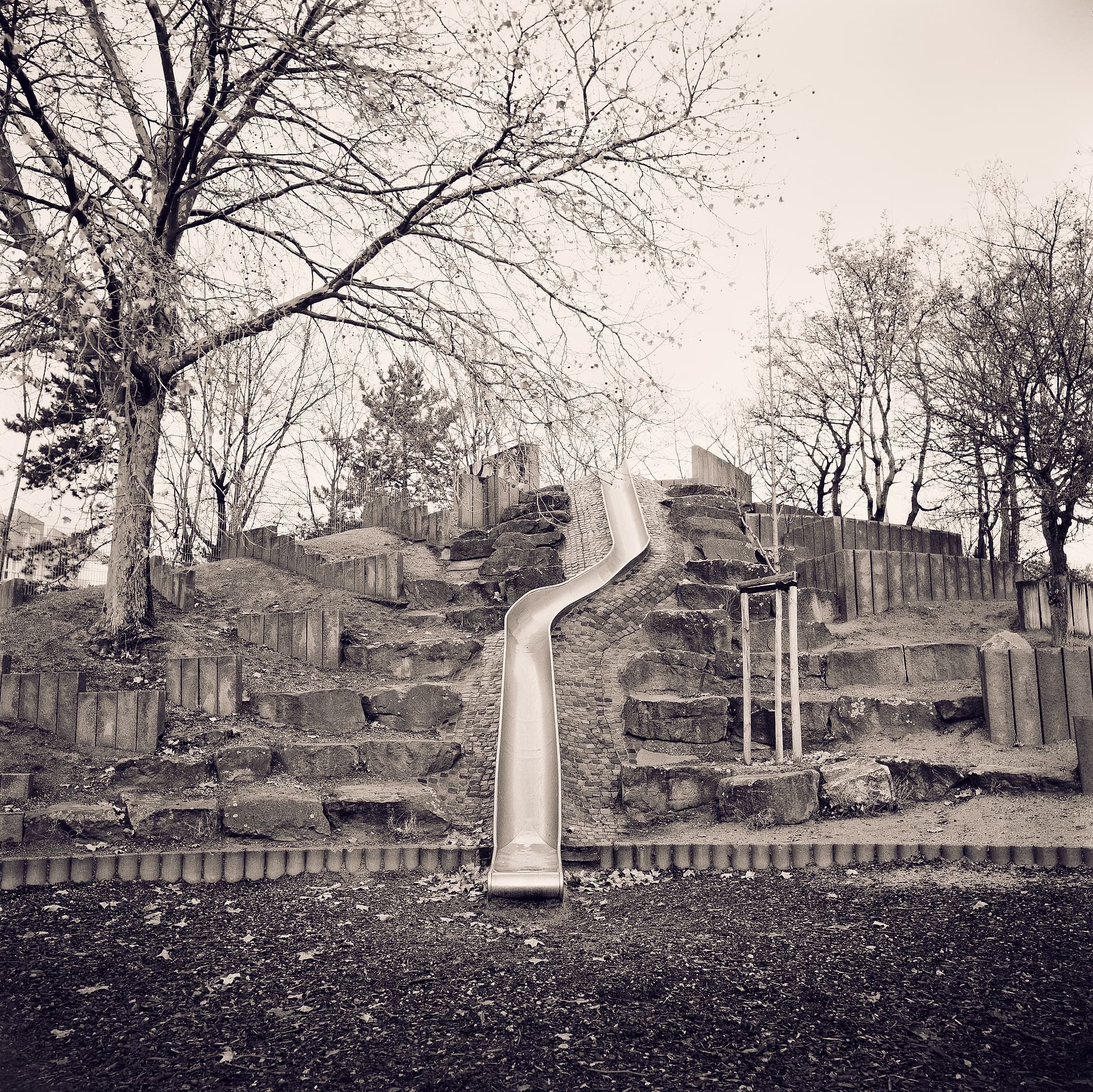 Metal slide running down a terraced stone and timber playground mound below bare trees and pale sky