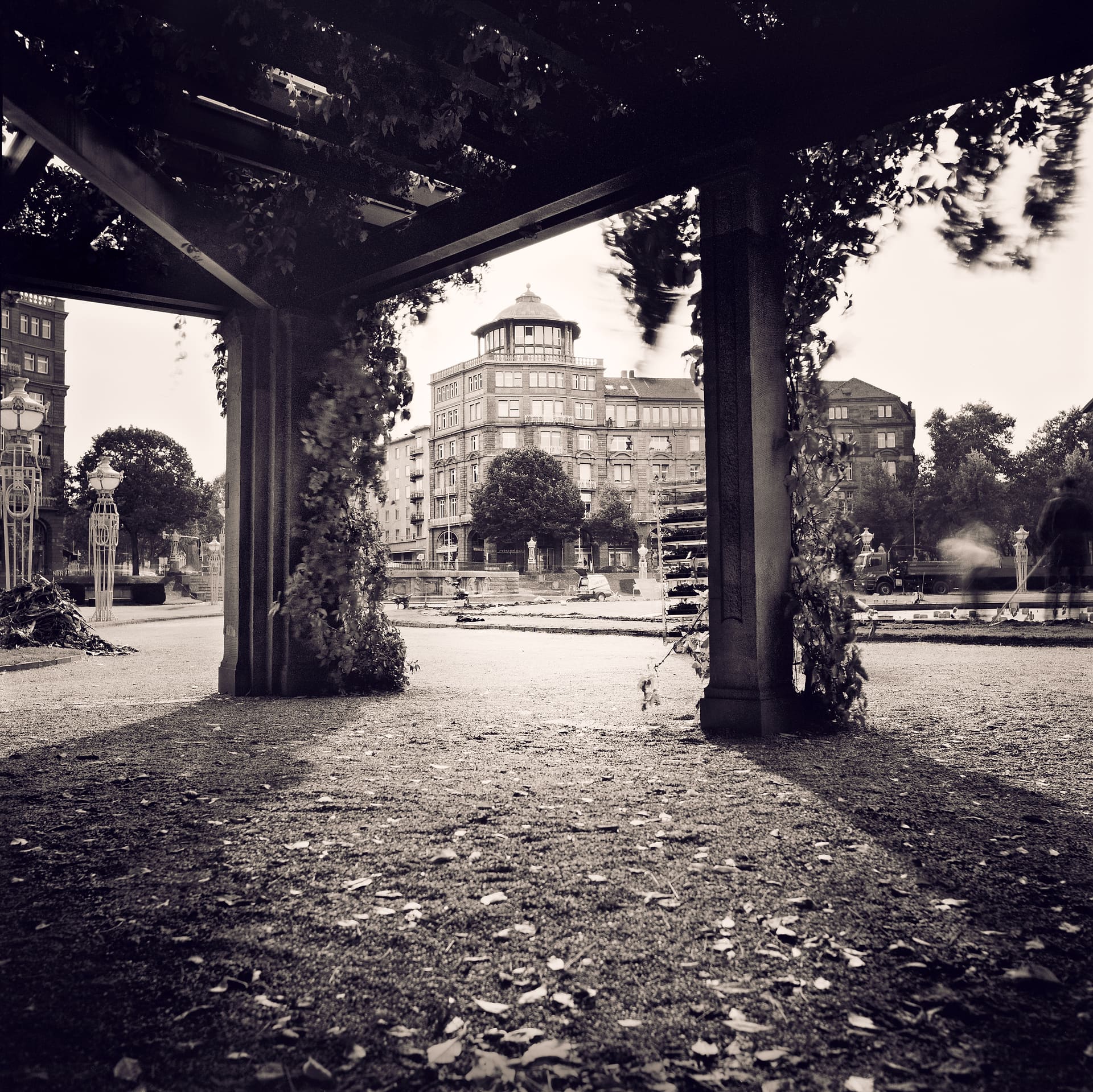 View from under a vine-covered pergola across gravel to a domed corner apartment block and ornate street lamps