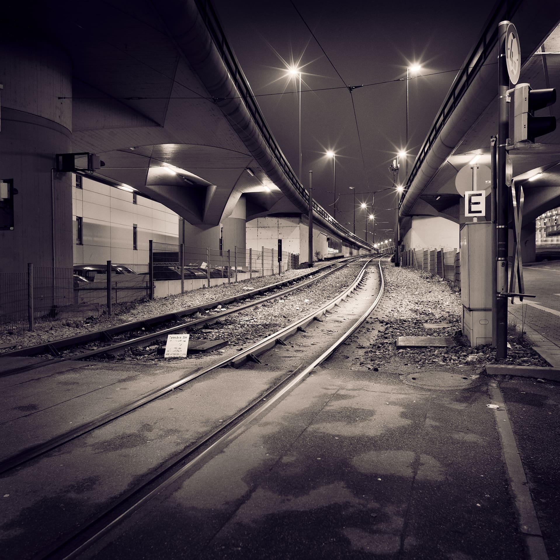 Tram rails curving beneath concrete flyovers at night, streetlamps flaring into stars above wet asphalt