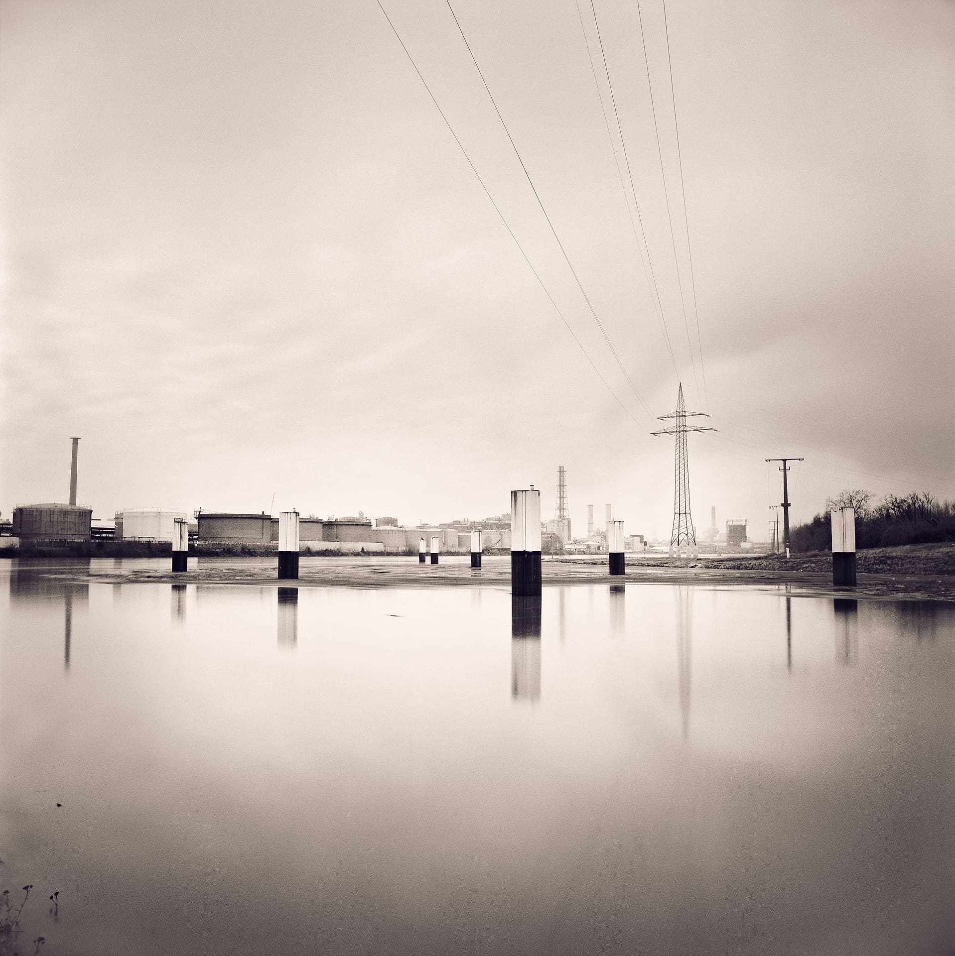 Chemical works, storage tanks and a pylon across glassy water, concrete stumps reflected under pale overcast sky