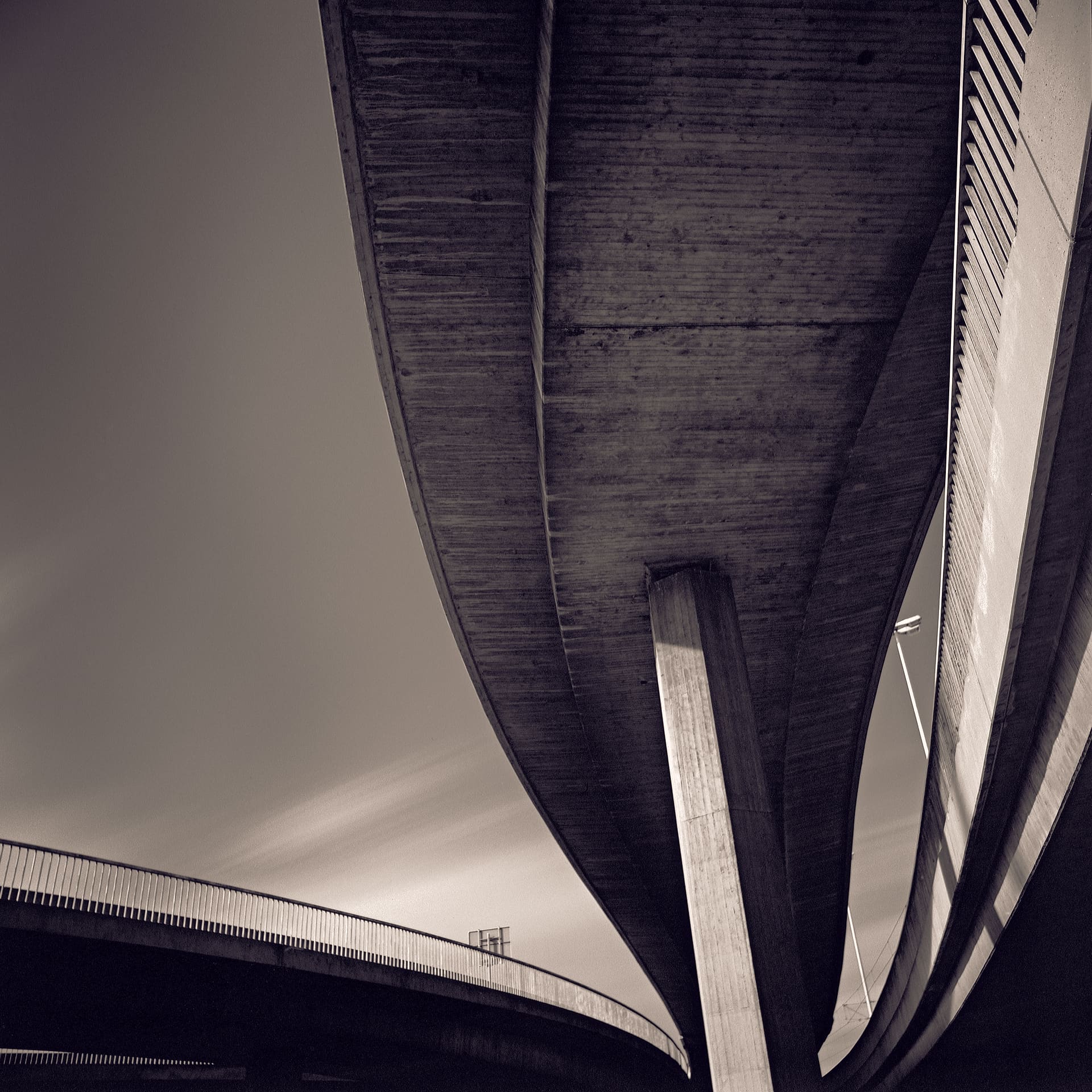 Underside of a curving concrete flyover meeting its single pier, second deck below, streaked cloud in dark sky