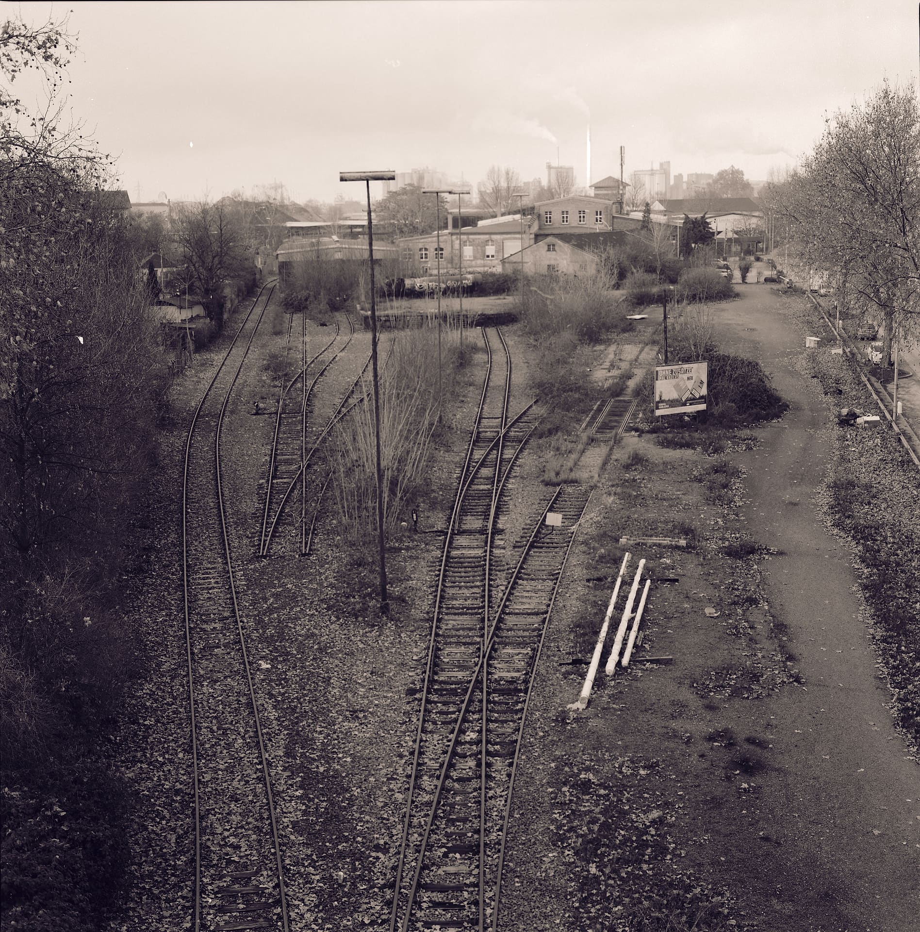 Disused railway sidings converging through weeds toward workshops and a smoking chimney under flat winter sky