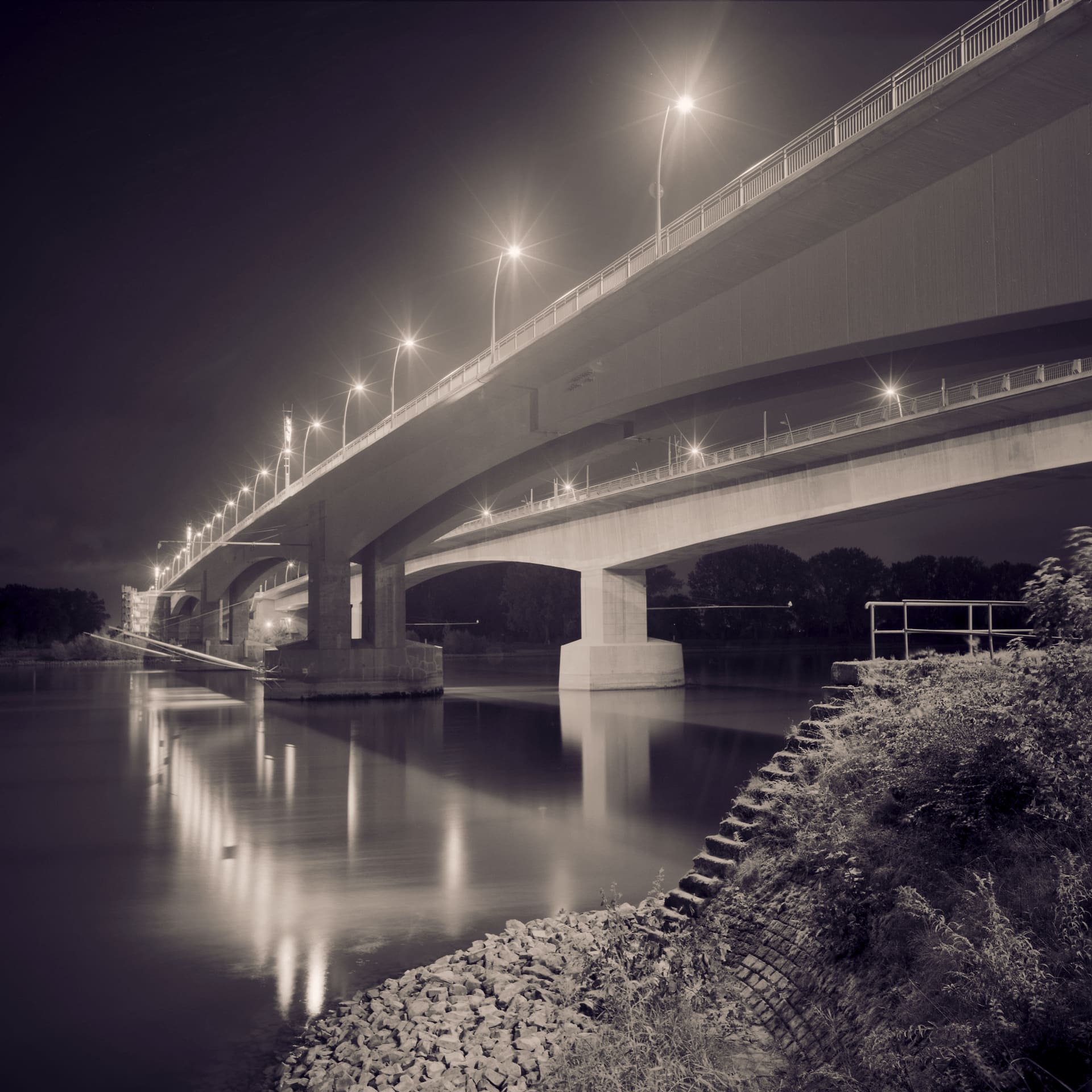 Twin concrete road bridges over a river at night, lamp reflections smeared down the smoothed water