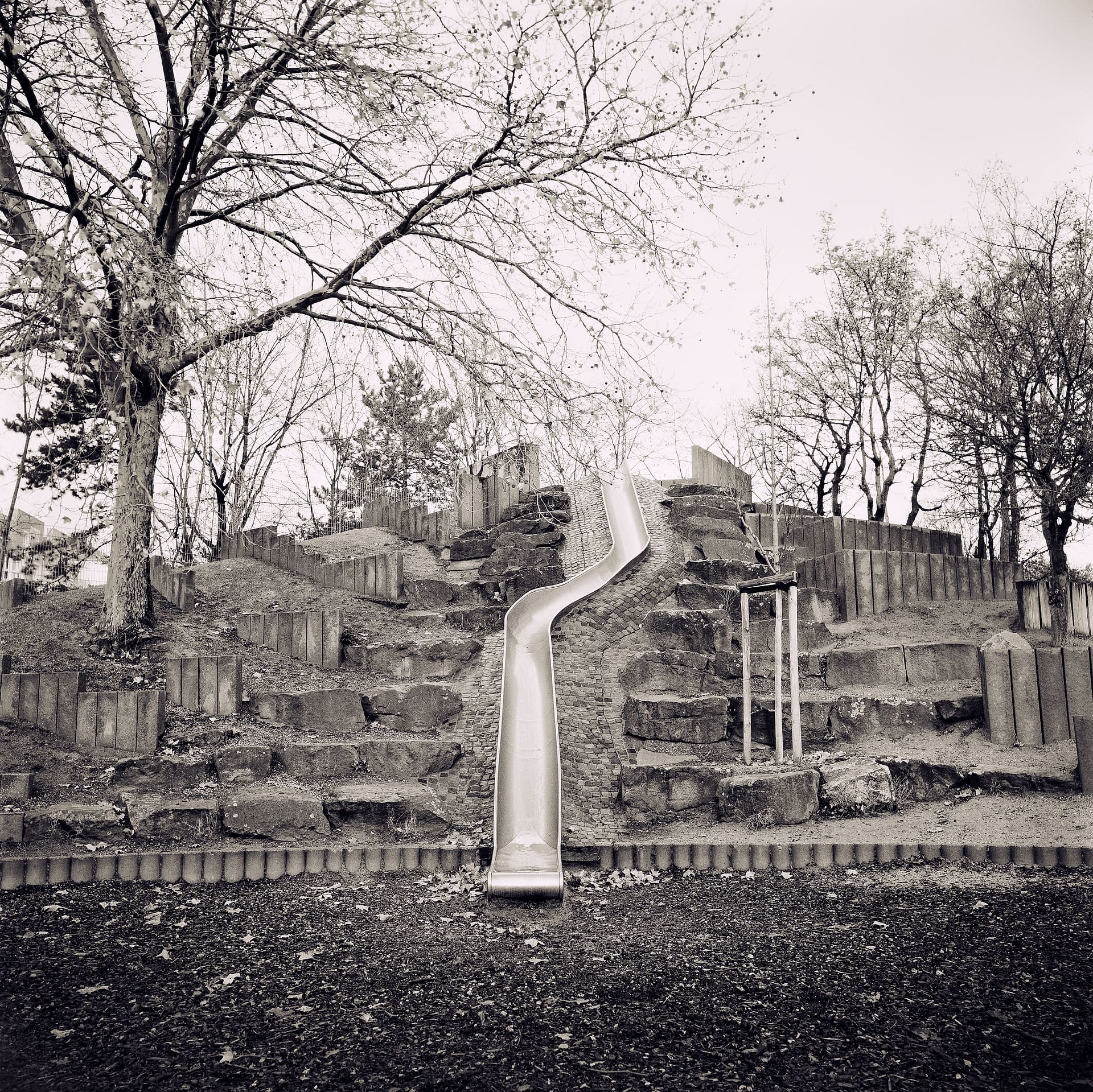 Metal slide running down a terraced stone and timber playground mound below bare trees and pale sky