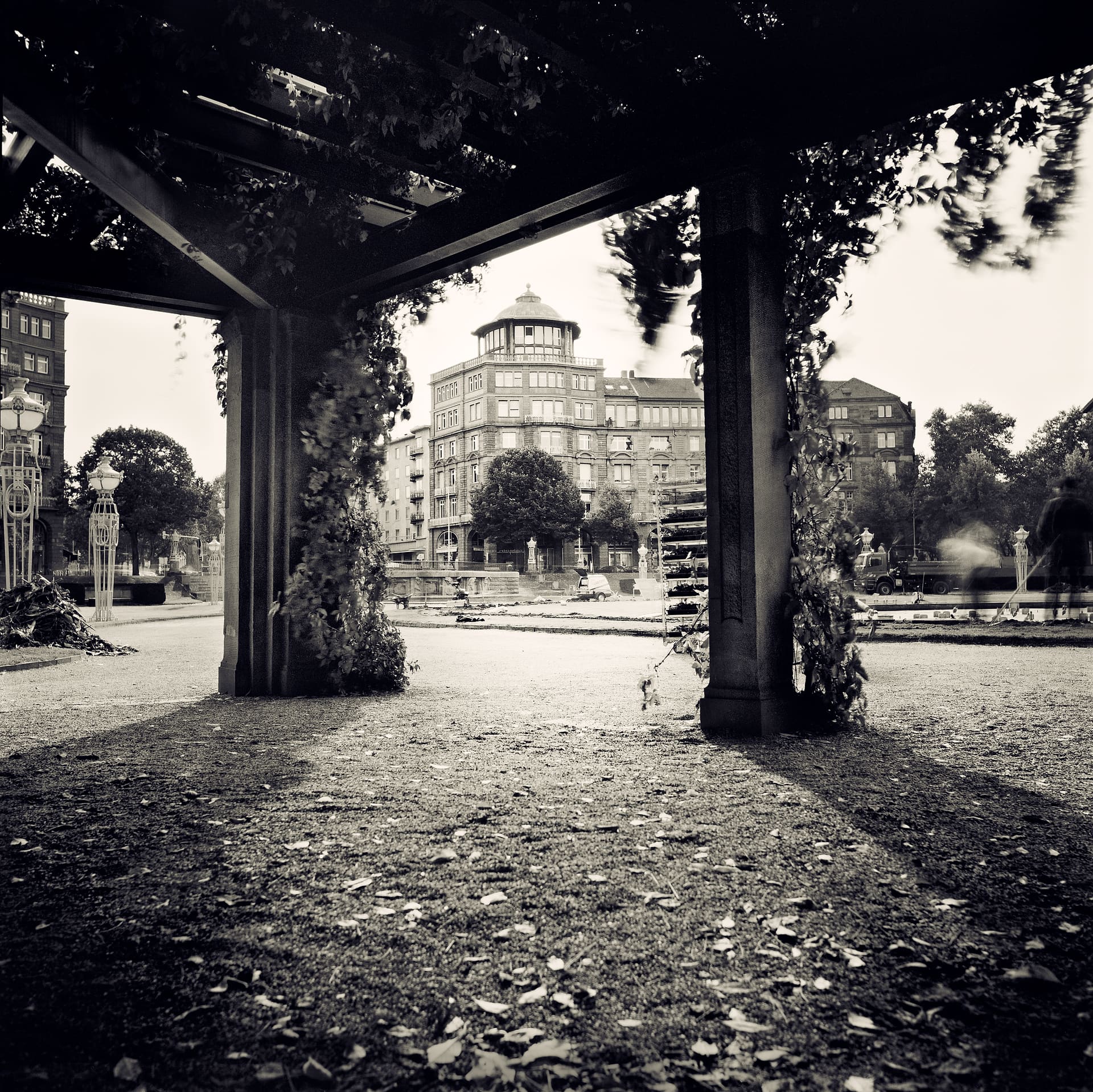 View from under a vine-covered pergola across gravel to a domed corner apartment block and ornate street lamps
