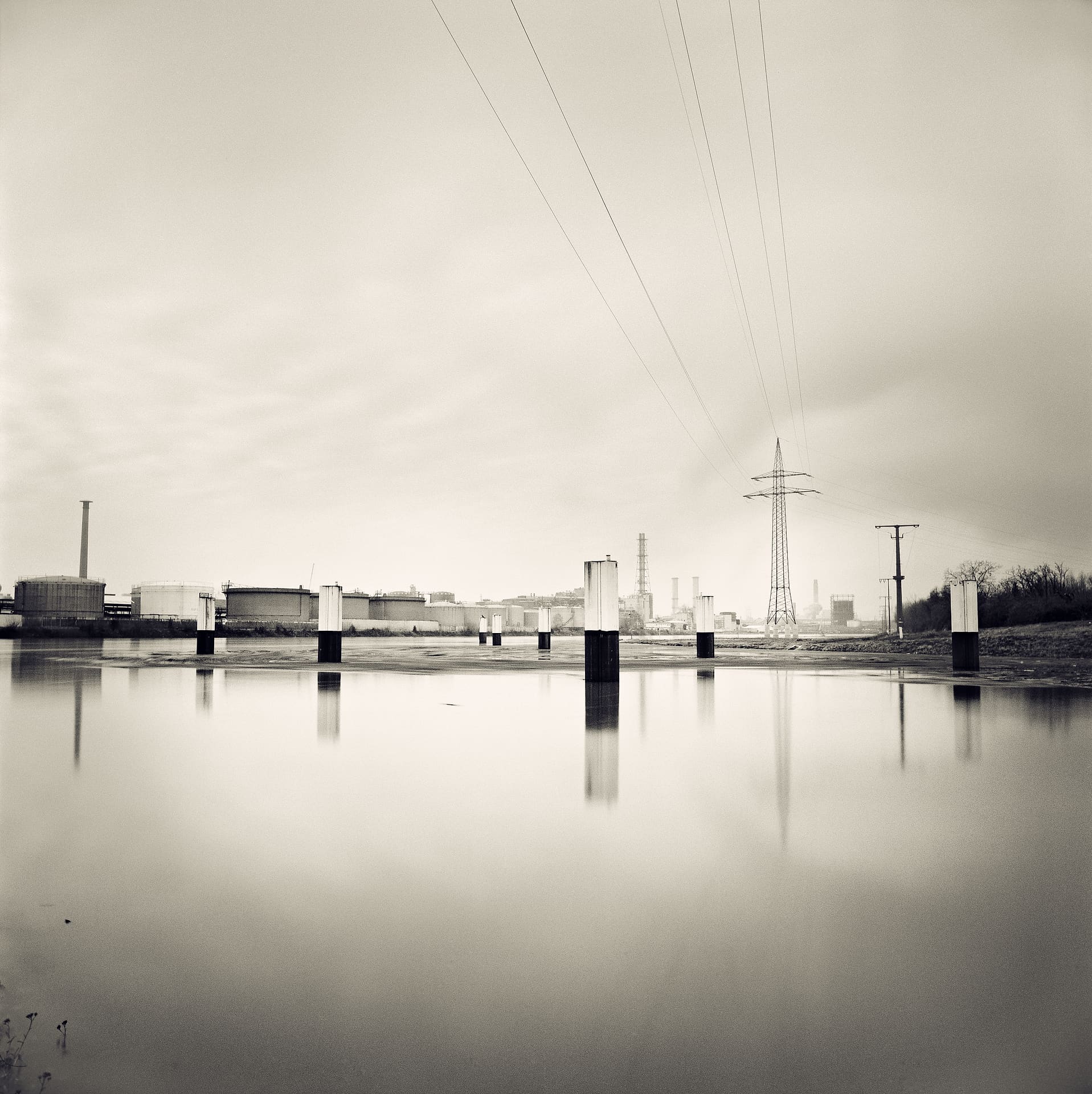 Chemical works, storage tanks and a pylon across glassy water, concrete stumps reflected under pale overcast sky