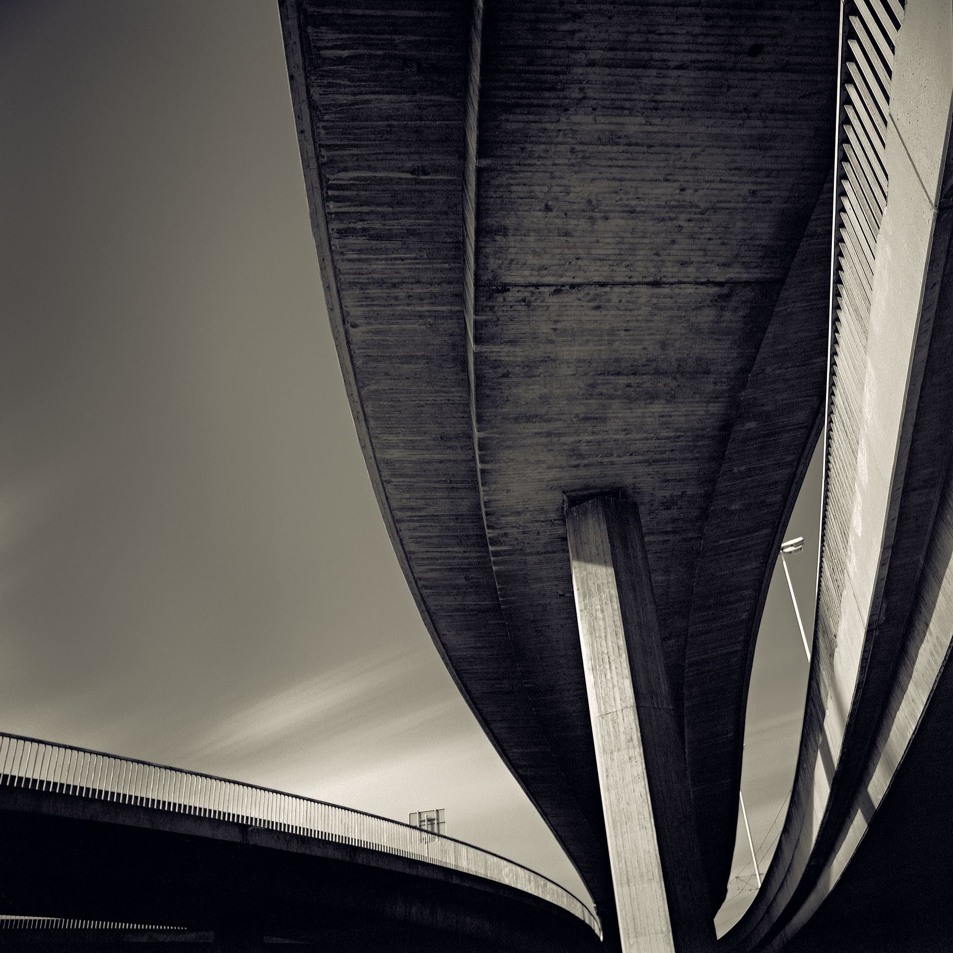 Underside of a curving concrete flyover meeting its single pier, second deck below, streaked cloud in dark sky