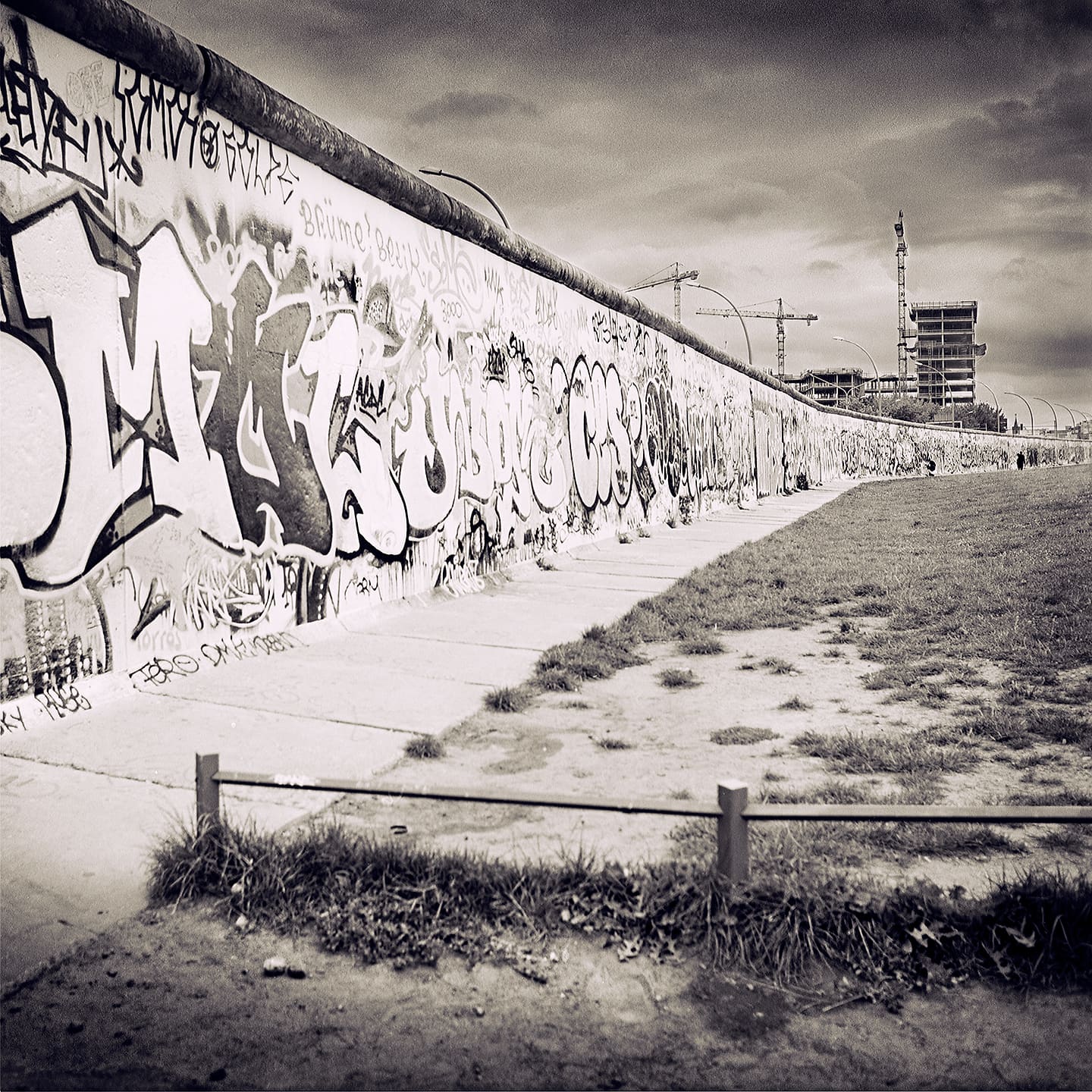 Graffiti-covered section of the Berlin Wall receding along a paved path, tower cranes over building work beyond