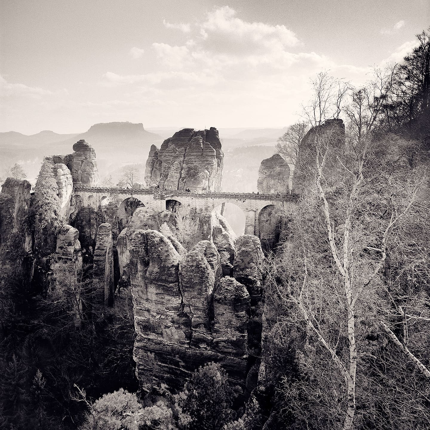 Stone arched bridge spanning between eroded sandstone pinnacles above a wooded valley, hazy hills beyond