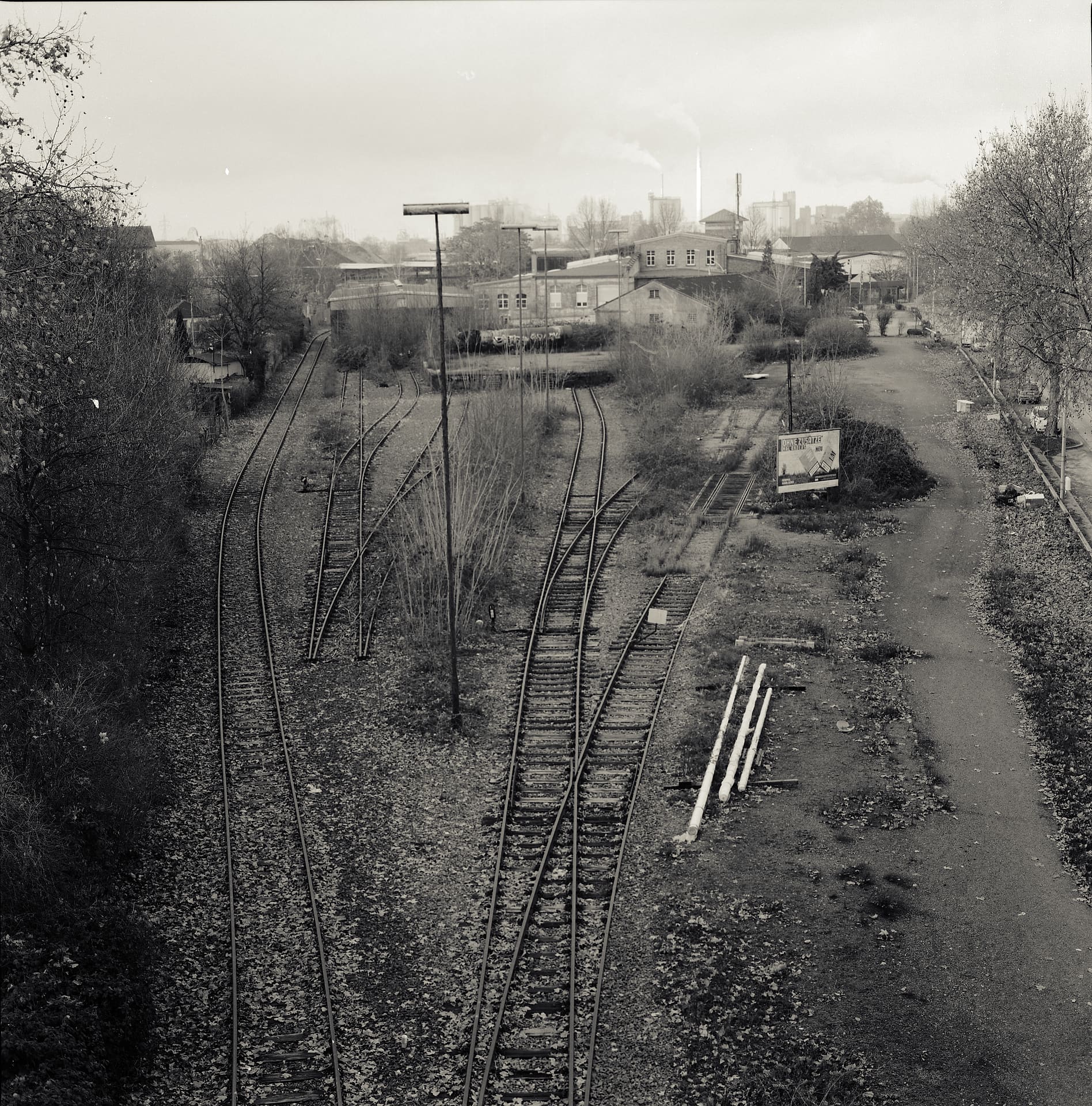Disused railway sidings converging through weeds toward workshops and a smoking chimney under flat winter sky