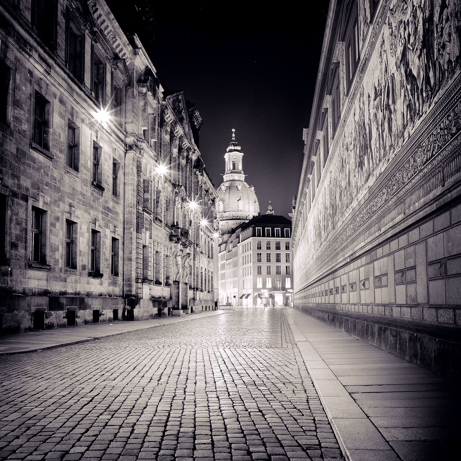 Empty cobbled street at night running toward the lit dome of the Frauenkirche, tiled Fürstenzug mural at right