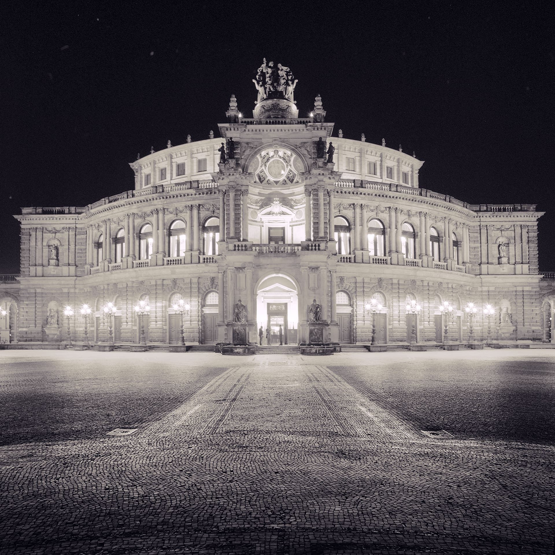 Floodlit neoclassical opera house with quadriga sculpture above its entrance, empty cobbled square in the foreground at night