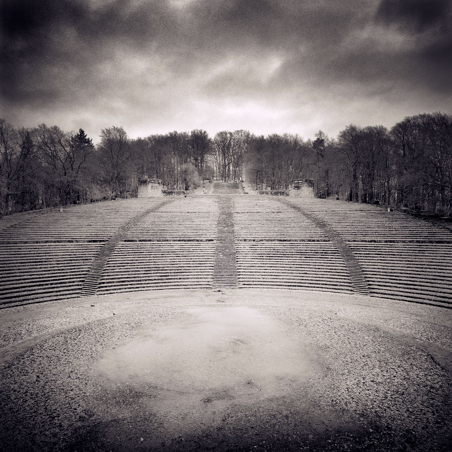 Empty open-air amphitheatre of stone terraces rising symmetrically to bare woods under heavy clouds