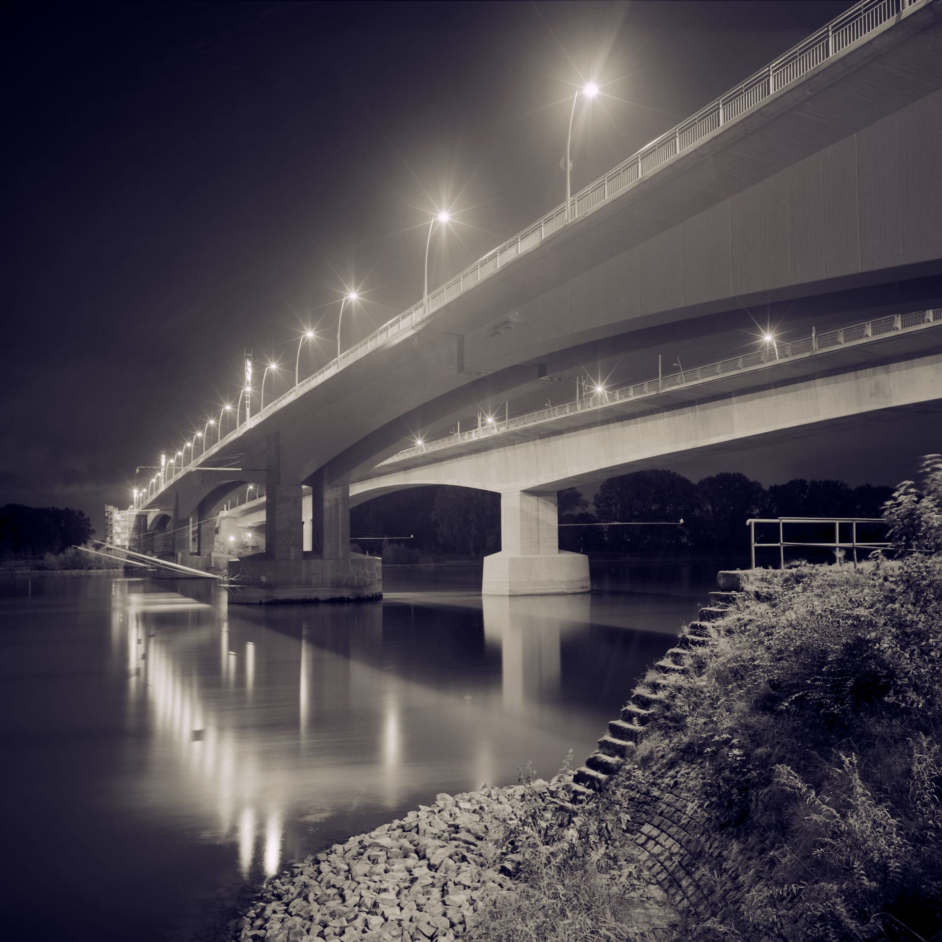 Twin concrete road bridges over a river at night, lamp reflections smeared down the smoothed water