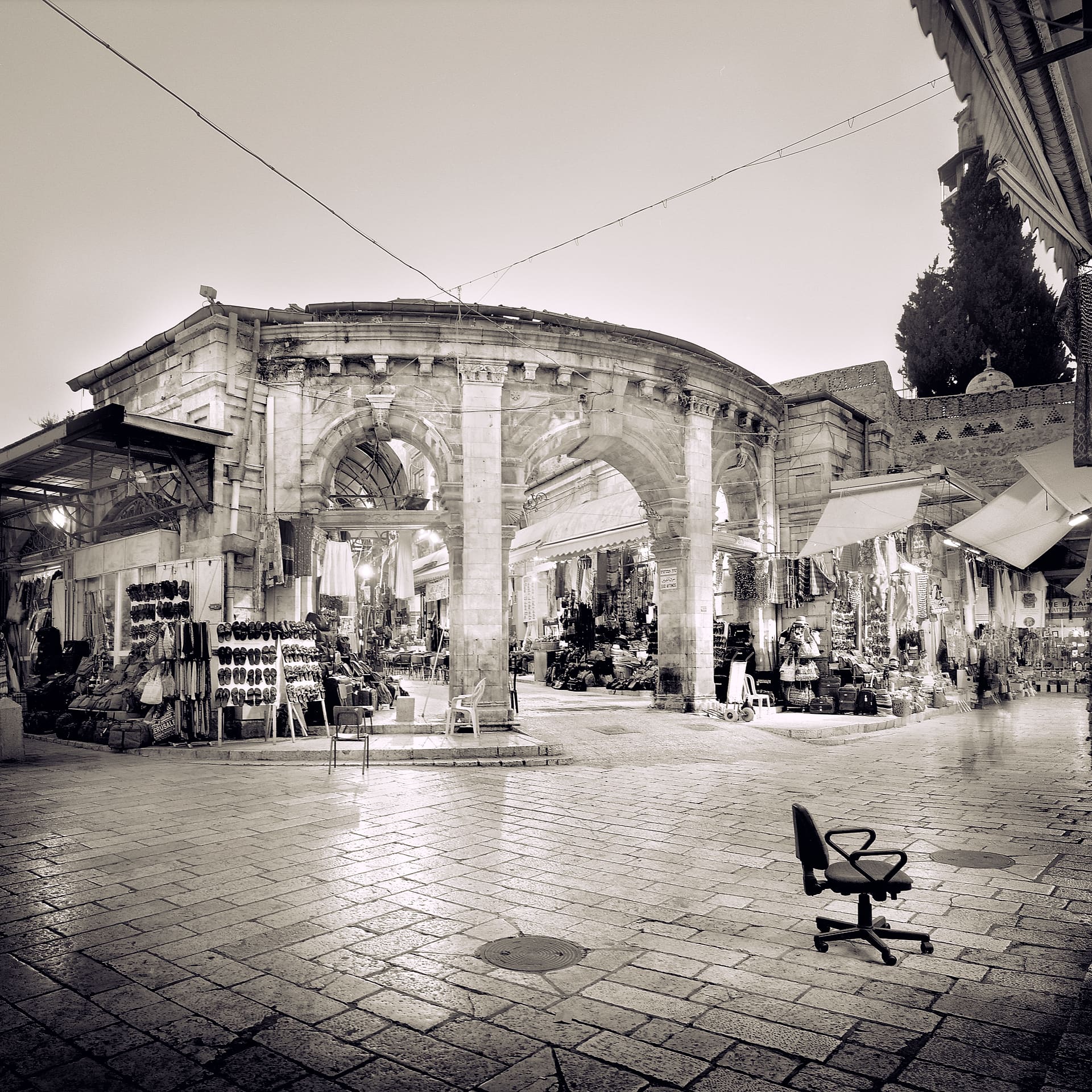 Lone office chair on wet flagstones before an arcaded stone market building hung with souvenir stalls at dusk