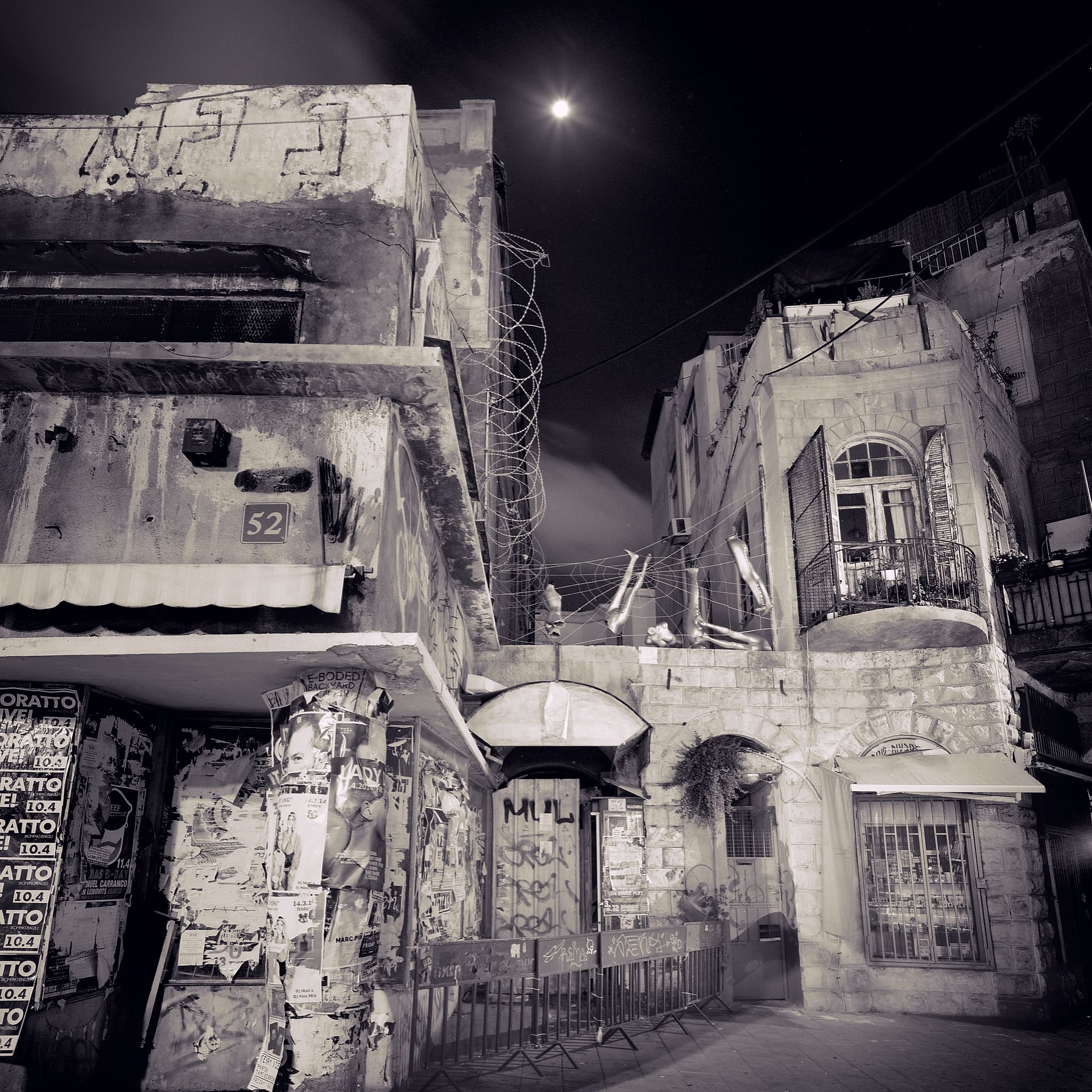 Moon above weathered street corner buildings at night, layered fly-posters, graffitied shutters and a wrought-iron balcony