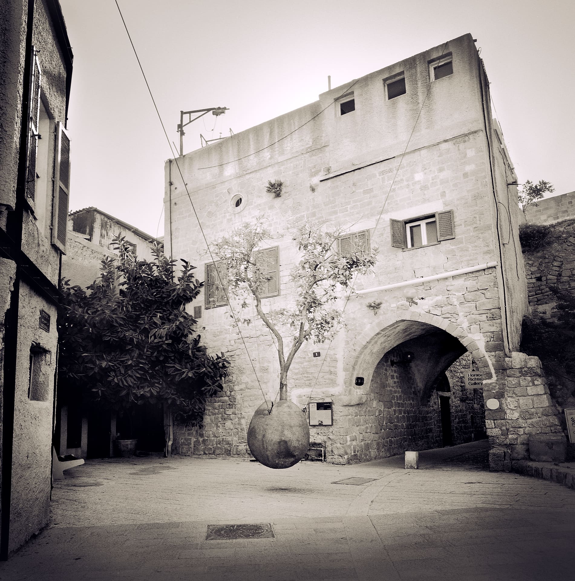 Small tree in an egg-shaped stone pot suspended by cables above an empty courtyard beside an arched stone building