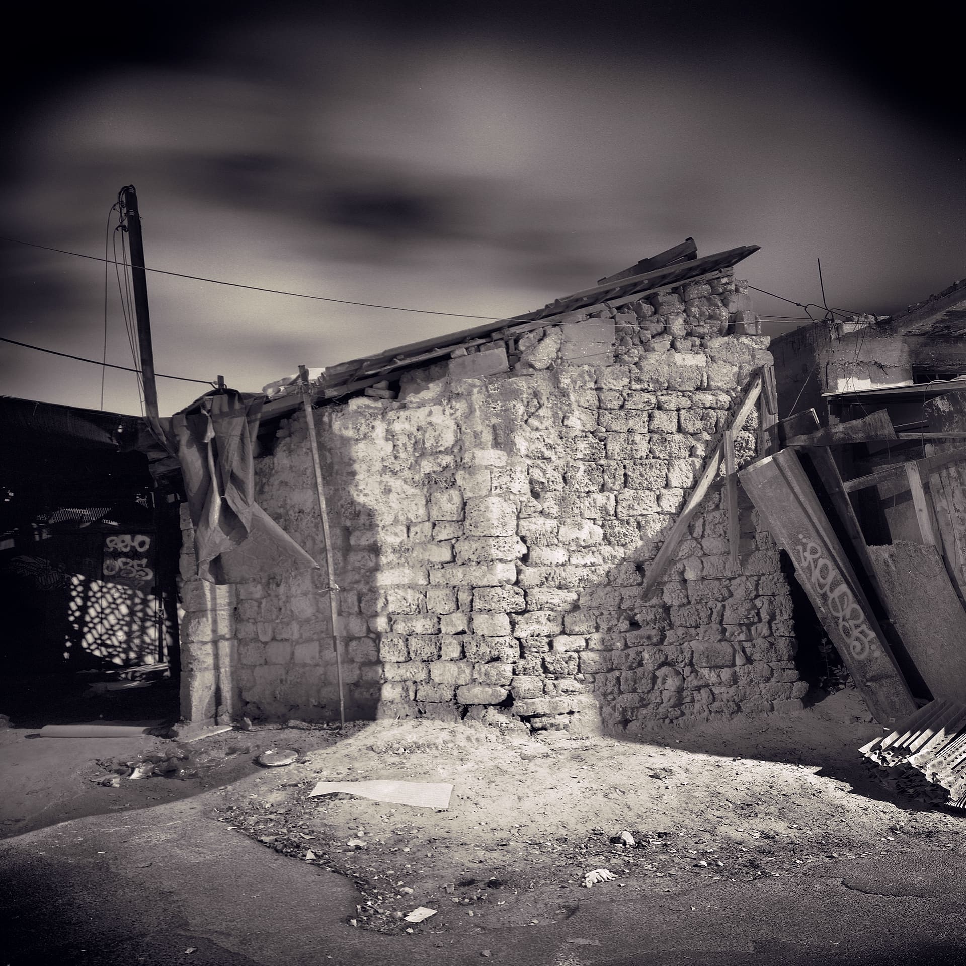 Roughly built stone wall with sagging tin roof and tarpaulin, rubble ground, streaked cloud in a long-exposure sky