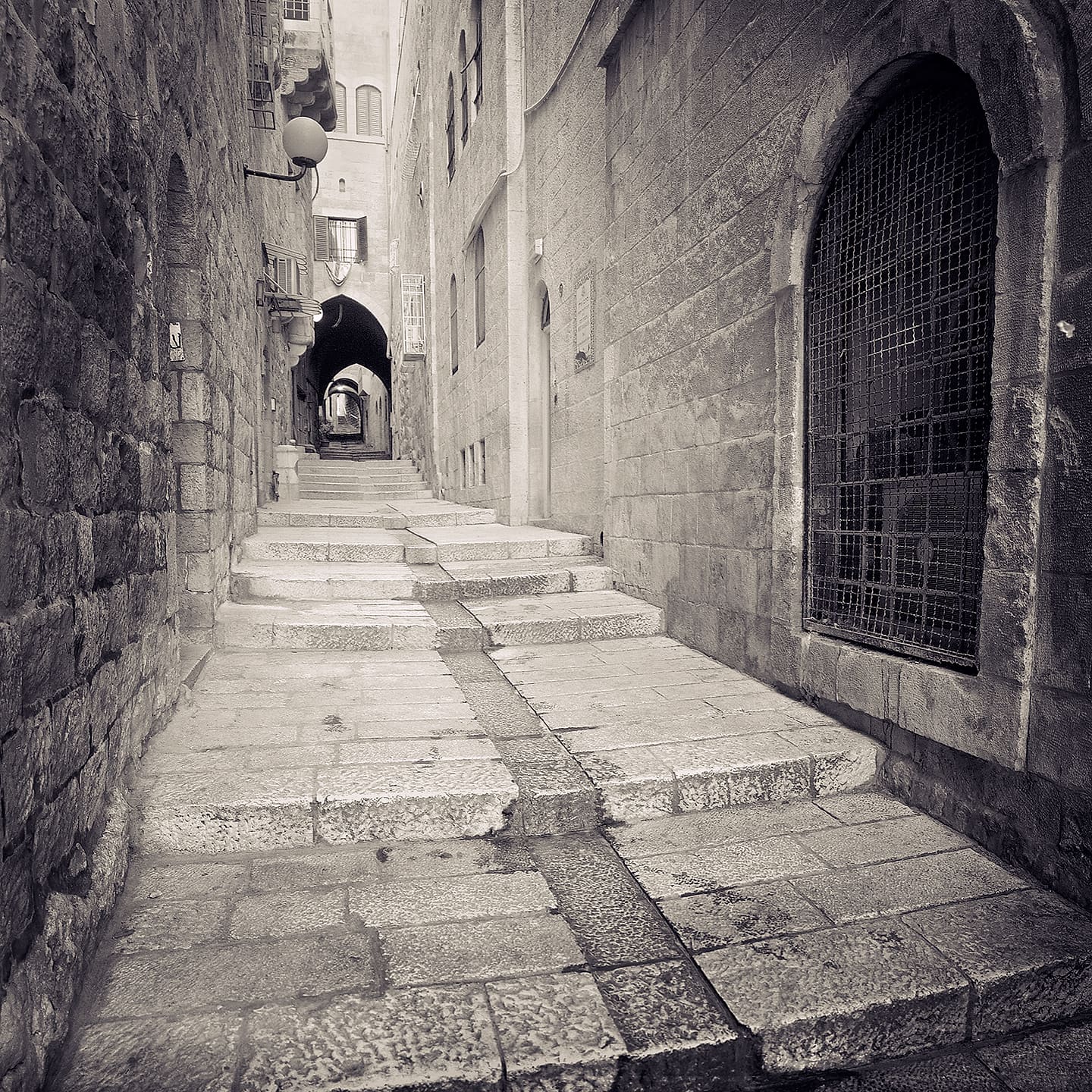 Empty stepped stone lane between tall walls, a grilled arched window at right and vaulted arch beyond