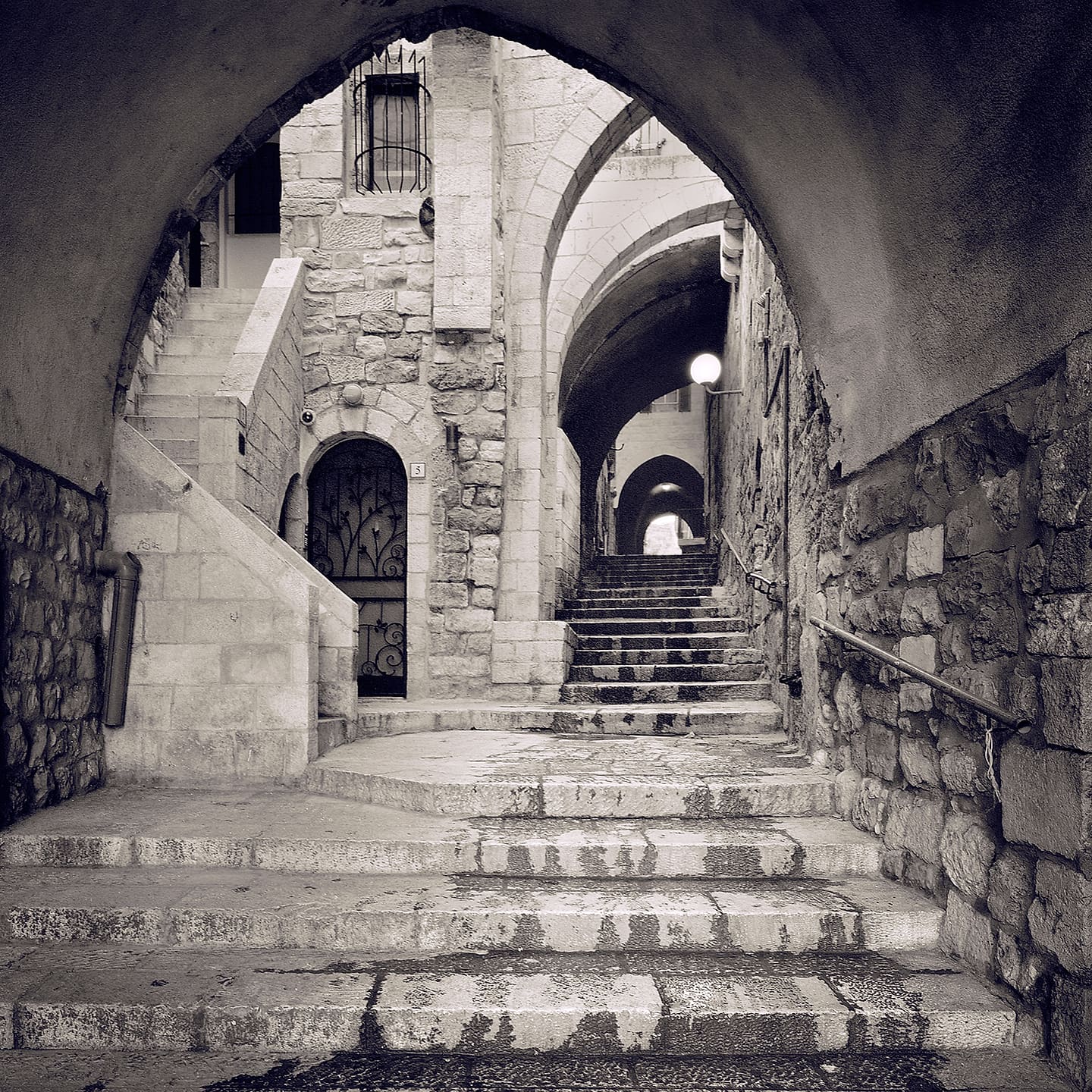 View through a stone arch to worn steps rising past an ironwork door under receding vaulted arches