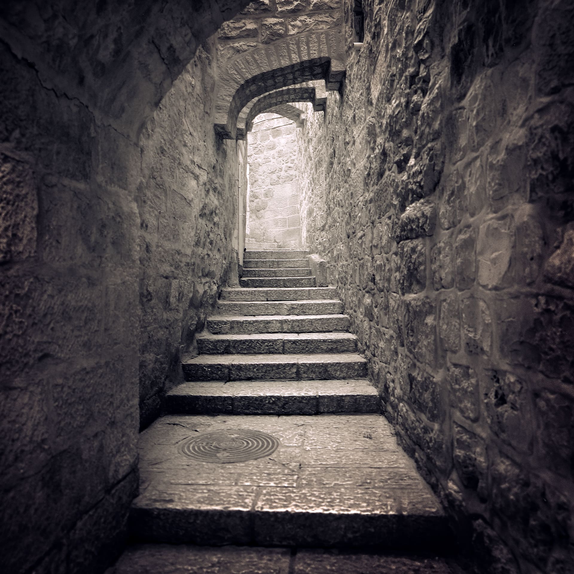 Narrow stone stairway climbing through a dim vaulted passage towards a brightly lit wall at the top