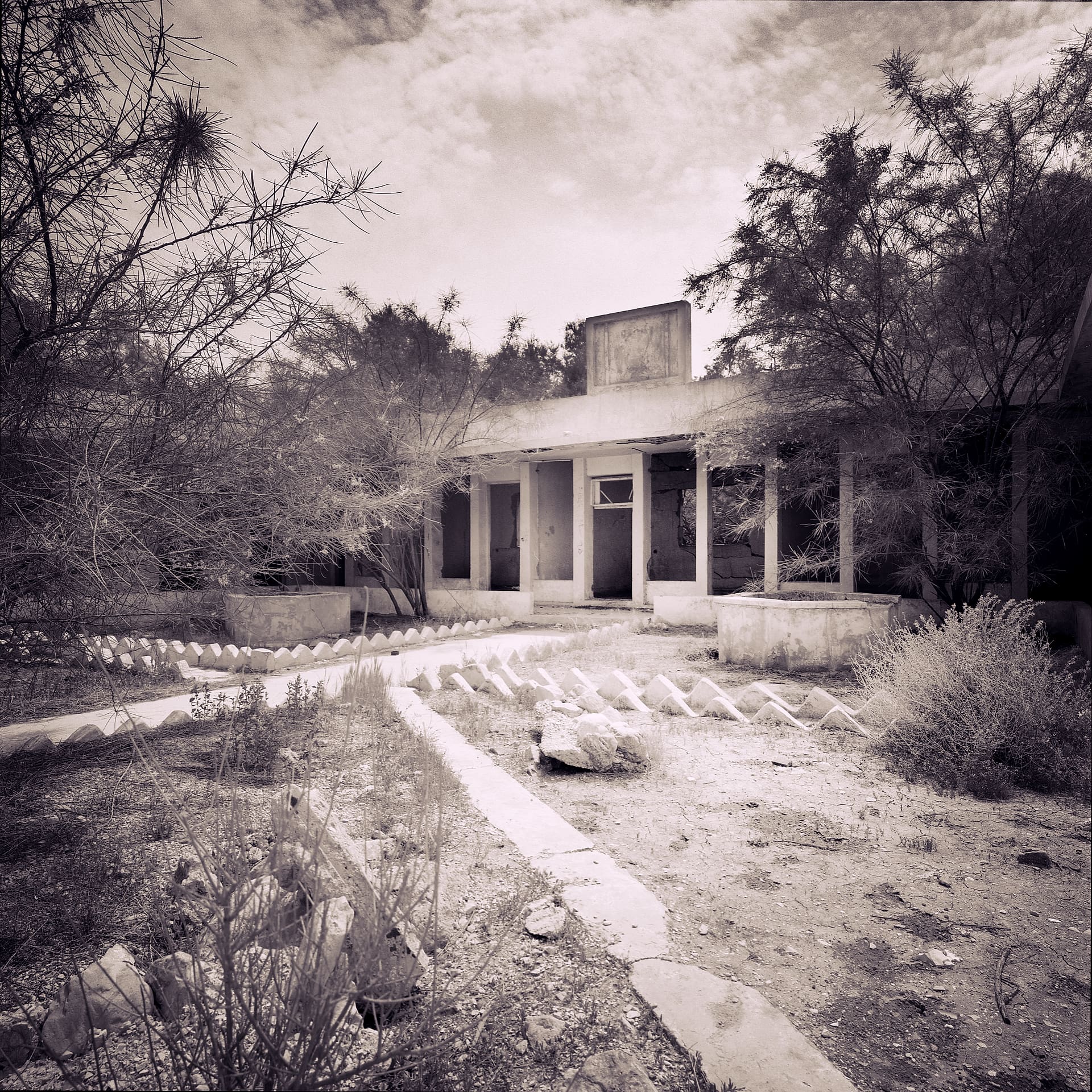 Abandoned flat-roofed concrete building with open colonnade, dry weeds and low kerbing overtaking its paved forecourt