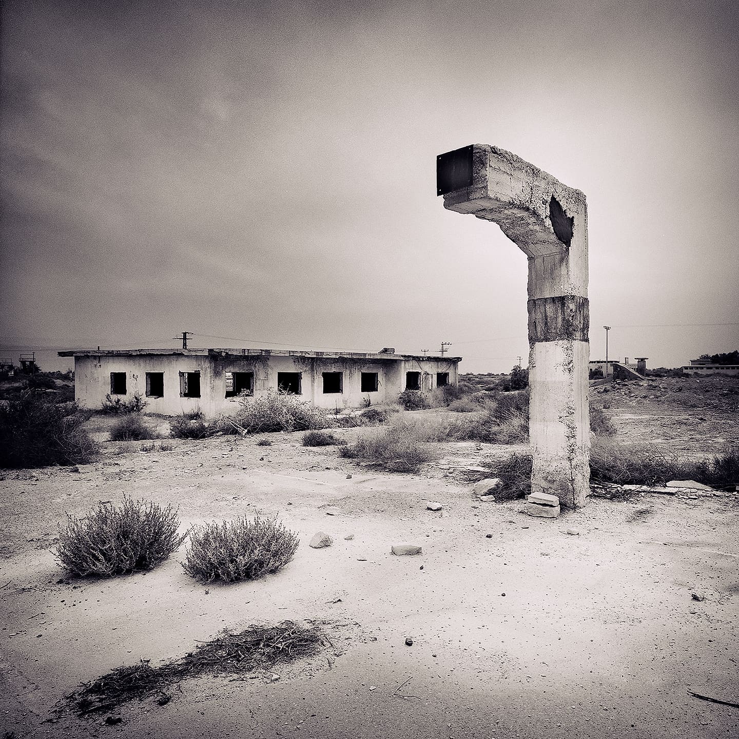 Free-standing concrete pillar with hooked crumbling top beside a derelict single-storey block of empty windows in sand