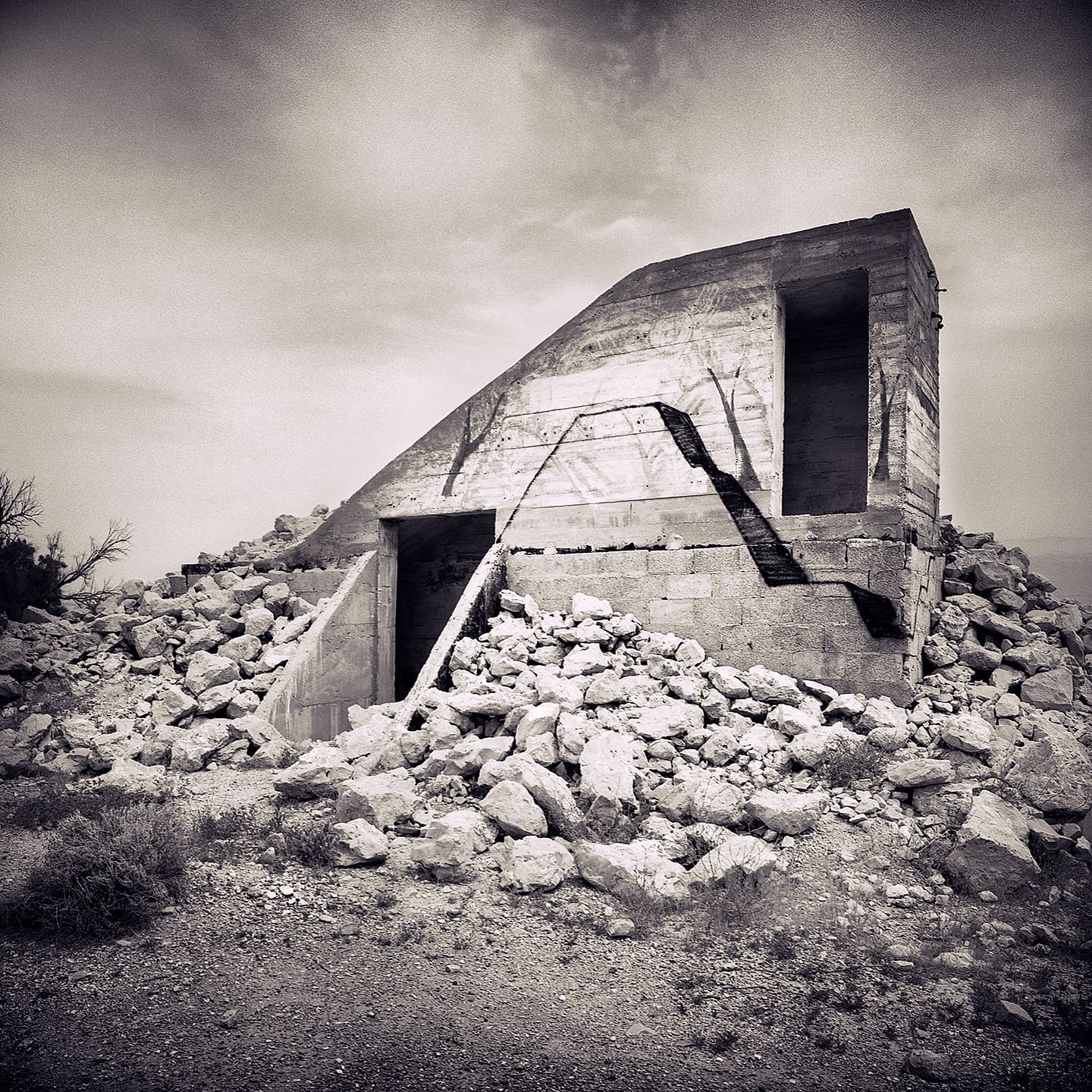 Wedge-shaped concrete shell with dark doorways, buried to its base in piled pale rubble under grey cloud