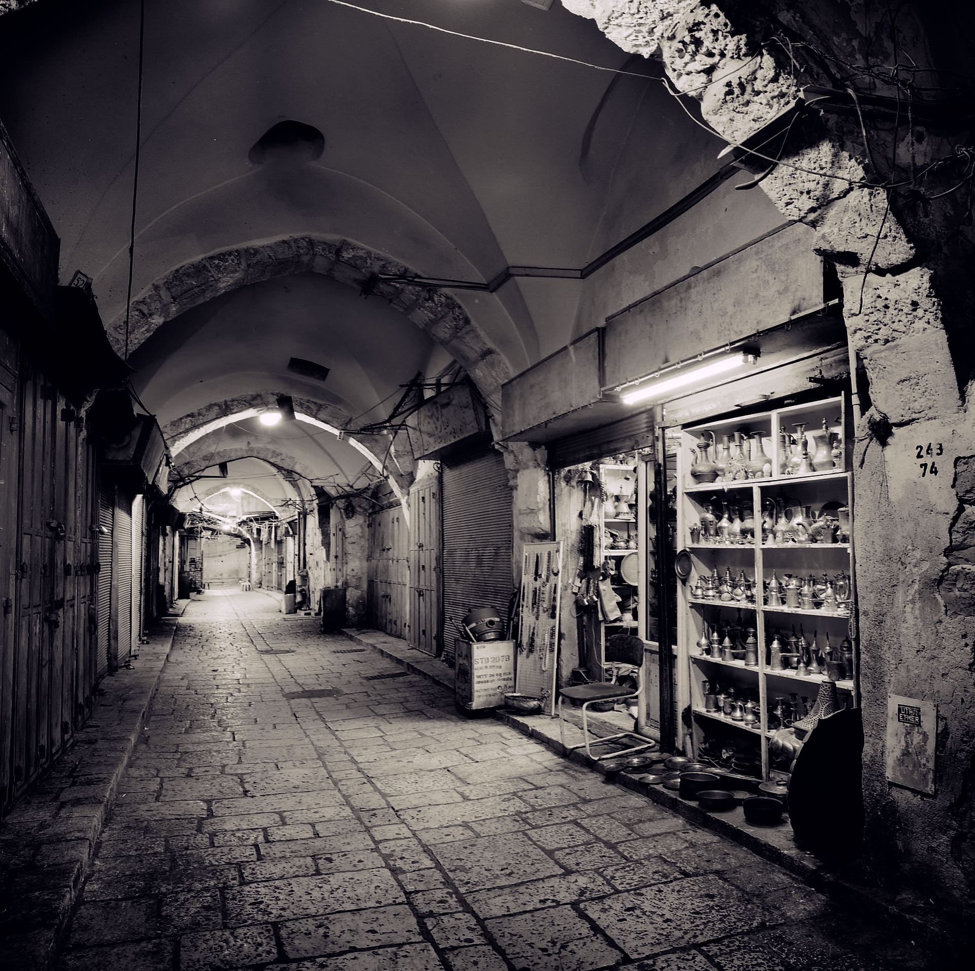 Vaulted covered bazaar at night, shutters closed except one lit shop stacked with brass and ceramic vessels