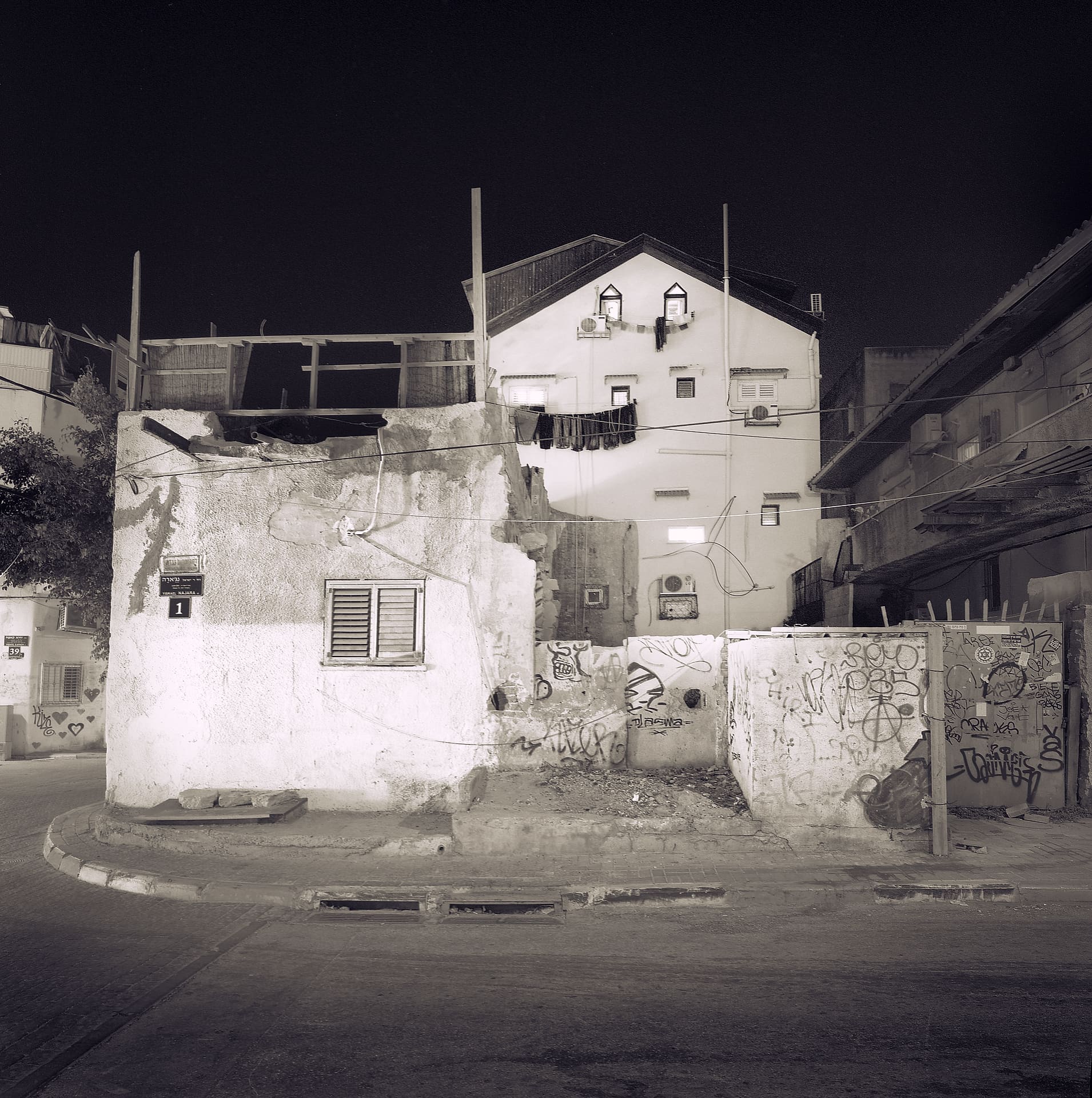 Squat whitewashed corner building with rooftop frame and graffiti walls, laundry strung on a taller house behind, at night