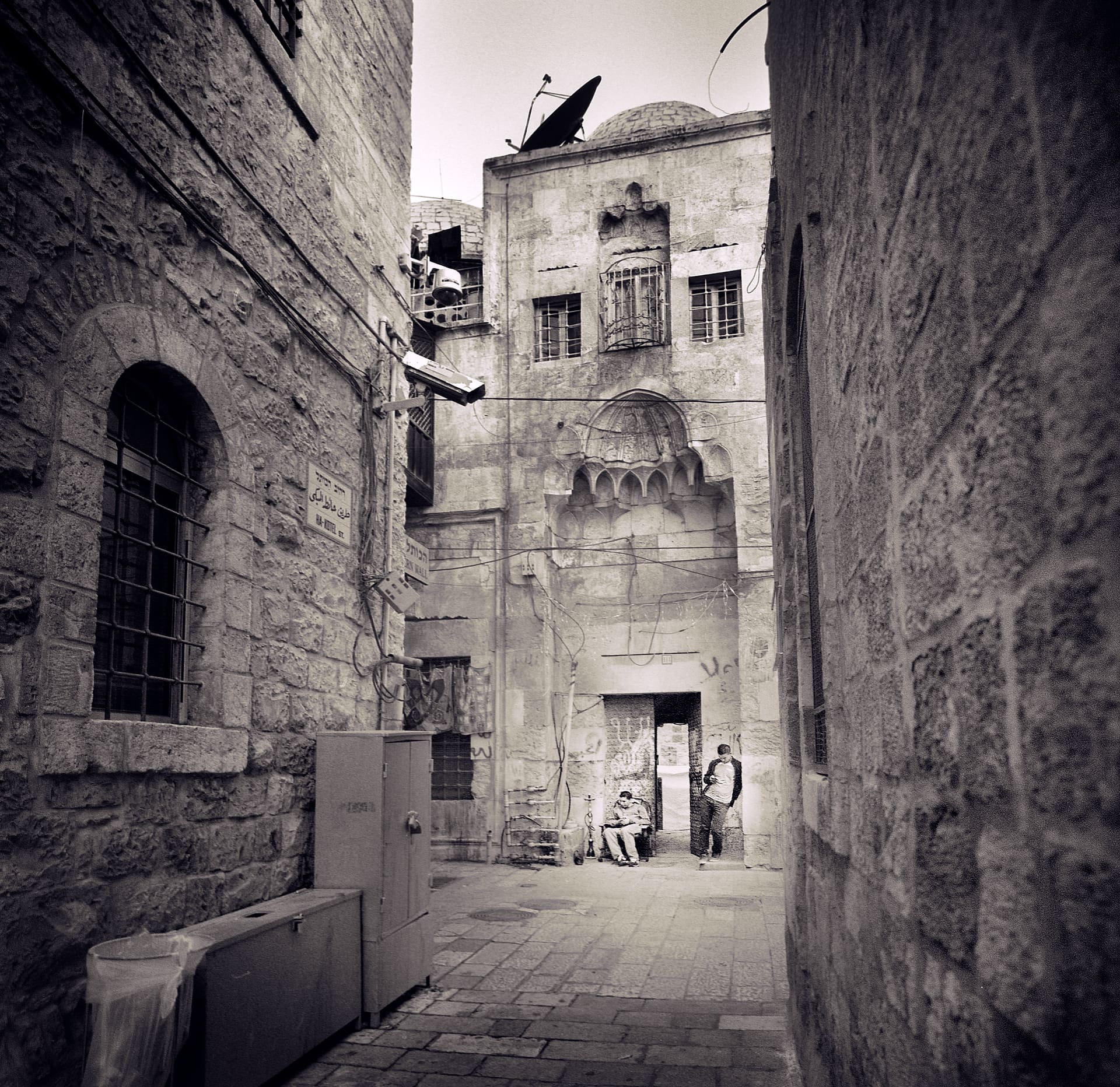 Narrow limestone alley opening onto an old stone building with satellite dish, dome and arched niche; two men rest below