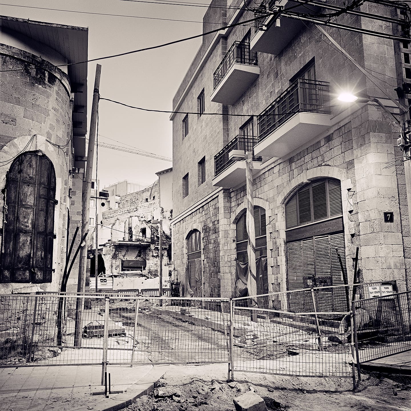 Stone apartment block with balconies beside a fenced-off demolition site and a lit streetlamp at dusk