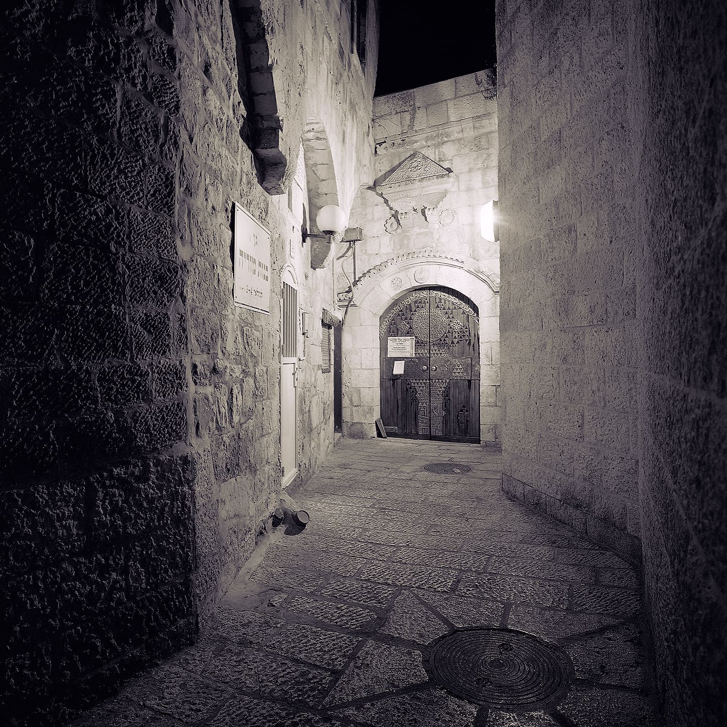 Narrow stone alley at night ending at a carved arched wooden door lit by wall lamps