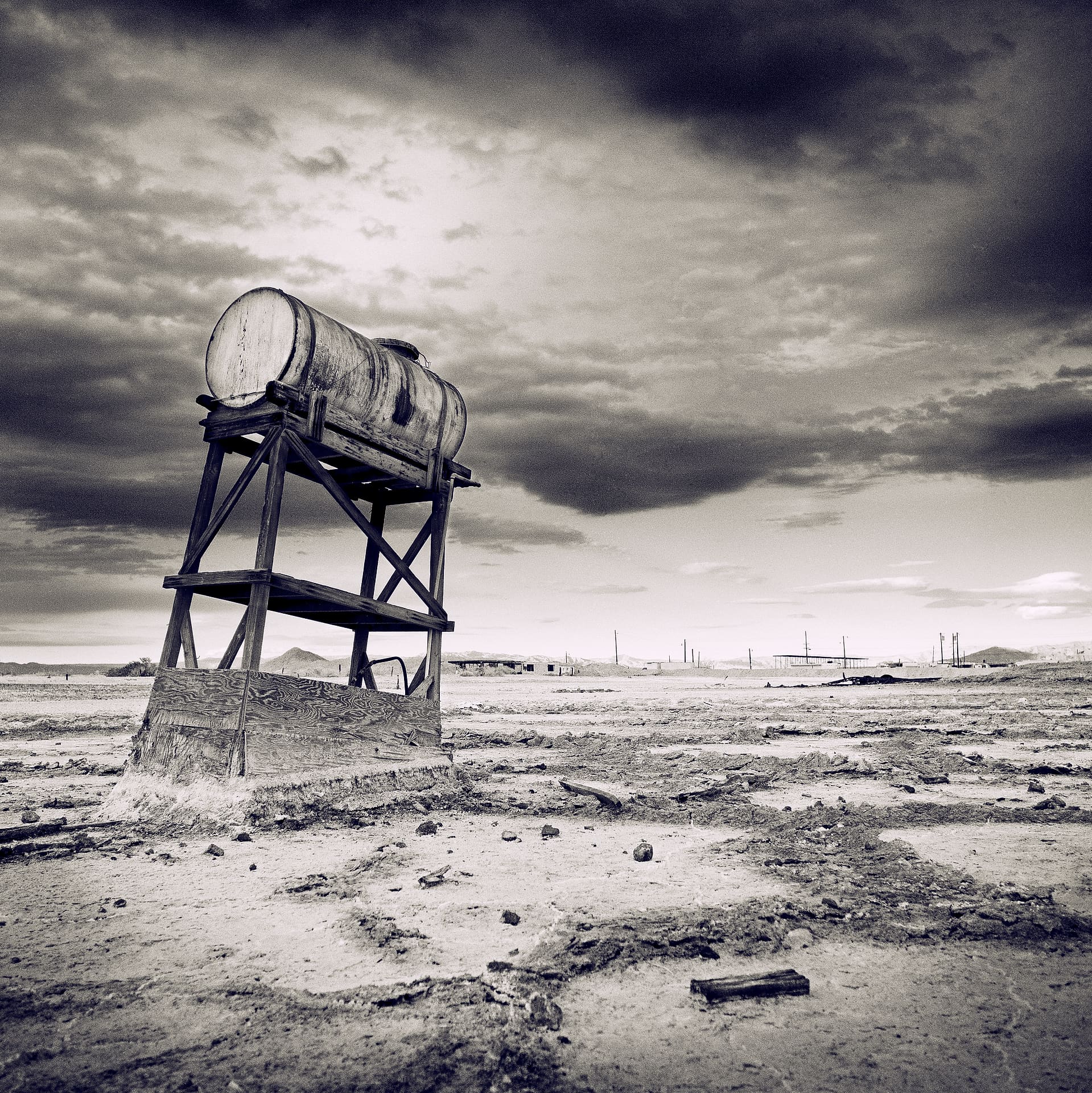 Leaning wooden tower carrying a cylindrical water tank on bare cracked ground beneath dark massed cloud